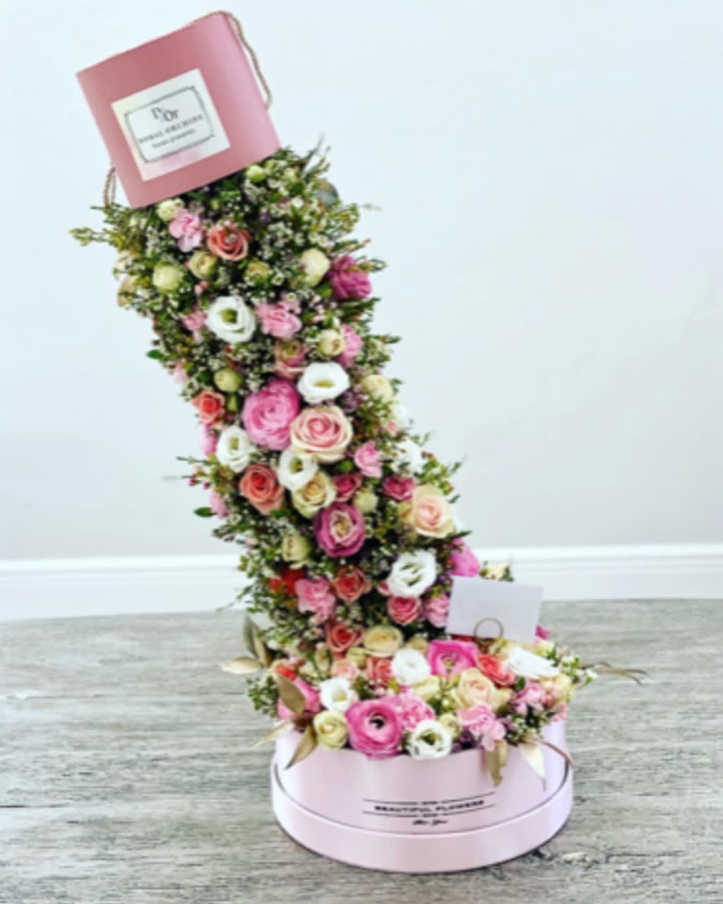 Irresistible Medium ™ - A breathtaking design where a pink hatbox tips forward, spilling out an abundance of roses, ranunculus, and seasonal blooms in pink and ivory tones. This “spilling garden” effect creates a whimsical, magical display.