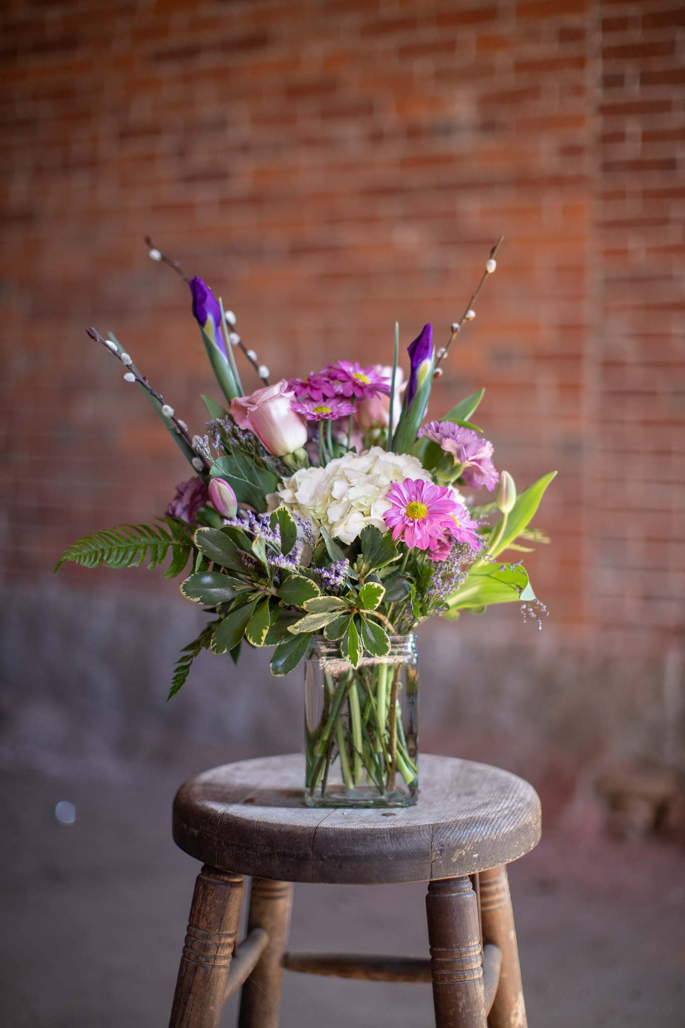 Iris and Floret - Beautiful budding iris blooms emerge from garden flowers arranged in a rectangular glass vase. Pretty hydrangea blooms with playful daisies, tulips and assorted foliage come together gracefully in this garden design. Branch material will vary based on season. Pussy willow is shown in photo, birch, snowberry, or curly willow will be used once season shifts. Colors may vary slightly on major floral holidays. Original design by Katie.