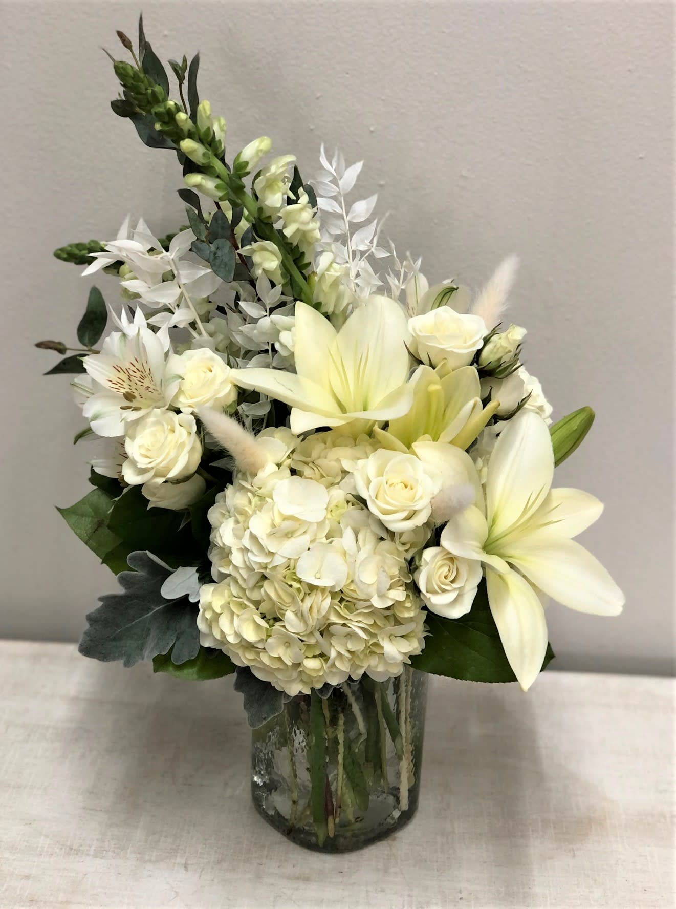 Trendy In White Bouquet - Mixture of white flowers such as lilies, spray roses, alstromeria, hydrangeas, snapdragon, gunni, dusty miller, dried ruscus, bunny tails arranged in a clear vase
