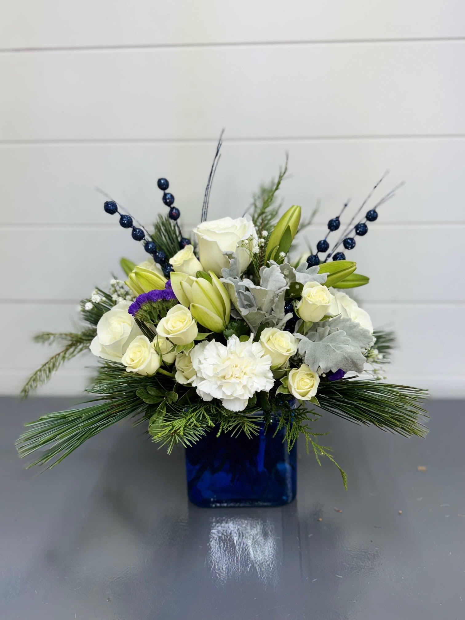 Sapphire &amp; Sparkles Bouquet - Sapphire &amp; Sparkles Bouquet Add a touch of elegance and festive charm with the Sapphire &amp; Sparkles Bouquet. This stunning arrangement features crisp white blooms accented with delicate purple statice, vibrant sparkly blue berries, and is presented in a stylish blue cube vase. Perfect for holiday gifting, winter celebrations, or brightening any room with a shimmering seasonal touch.   FCF-W18-05