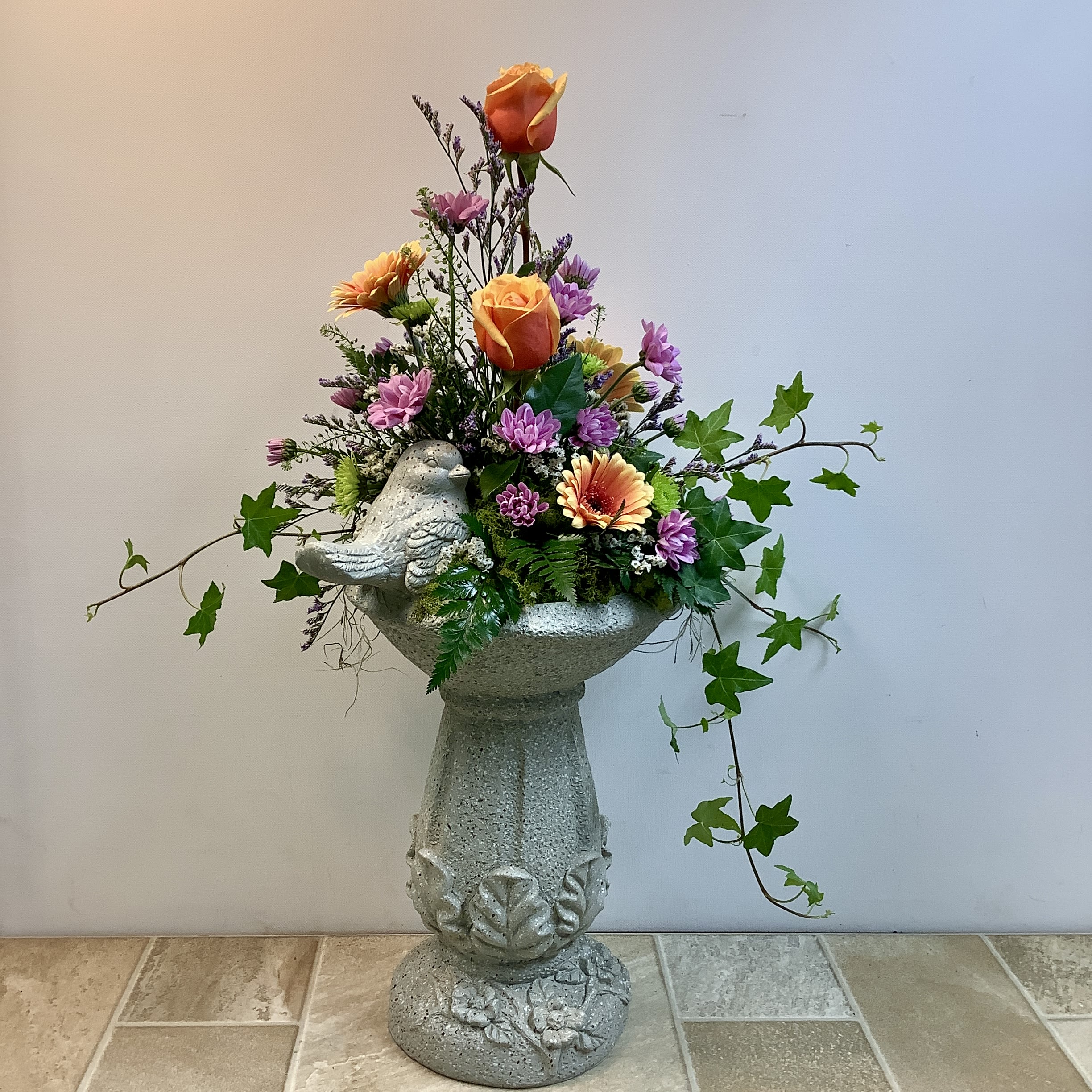 Larissa's Garden - This charming birdbath arrangement features a classic stone-look pedestal bowl adorned with a perched bird, creating a peaceful garden-inspired design. Rising from the center are vibrant cherry brandy roses that add height and warmth, surrounded by soft lavender and green cushion mums. Peach and coral gerbera daisies bring cheerful pops of color, while delicate filler flowers and lush greenery create fullness and texture. Cascading ivy trails gracefully over the sides, giving the piece a natural, freshly gathered garden feel. The combination of warm sunset tones and cool lavender hues makes this arrangement both lively and serene — perfect for sympathy, remembrance, or bringing a touch of the outdoors inside. It measures 19” wide x 28”. Tall.