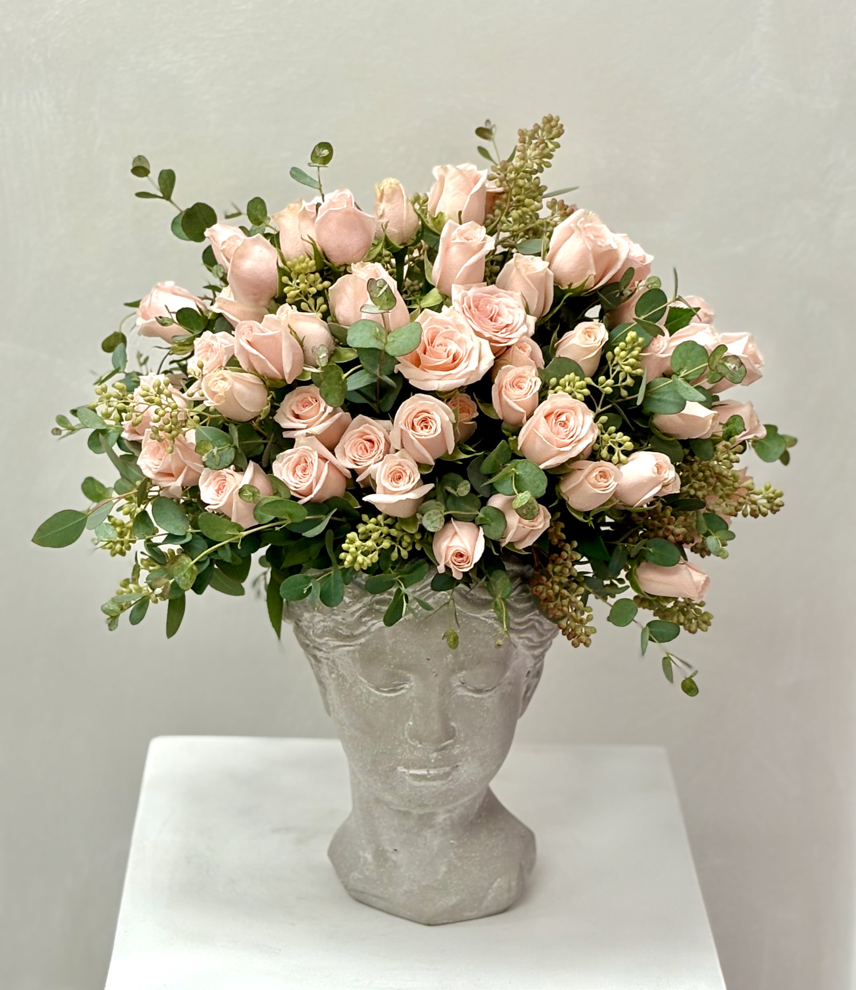 Blushing Muse By VLM Flowers - Soft blush spray roses bloom gracefully from a classical sculpted vase, accented with fresh eucalyptus and delicate seasonal greens. A refined, art-inspired arrangement that blends romance with timeless beauty.