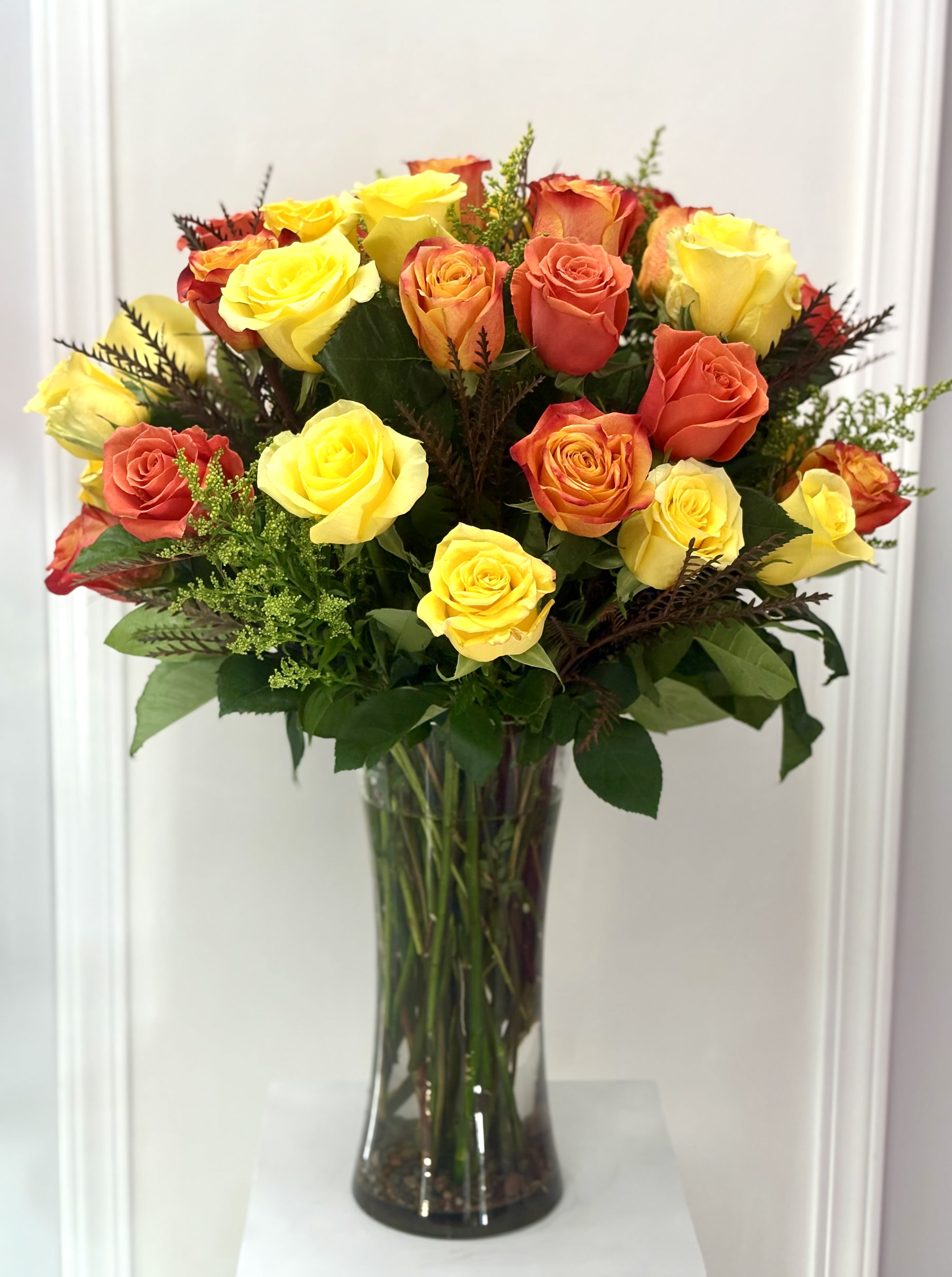 Sunset Radiance Vase By VLM Flowers - A vibrant display of yellow and orange roses arranged in a tall clear vase, inspired by the warm glow of a summer sunset. Accented with lush greenery, this cheerful bouquet brings brightness, energy, and joy to any space. Perfect for celebrations, birthdays, or uplifting someone’s day. In the photo deluxe size.