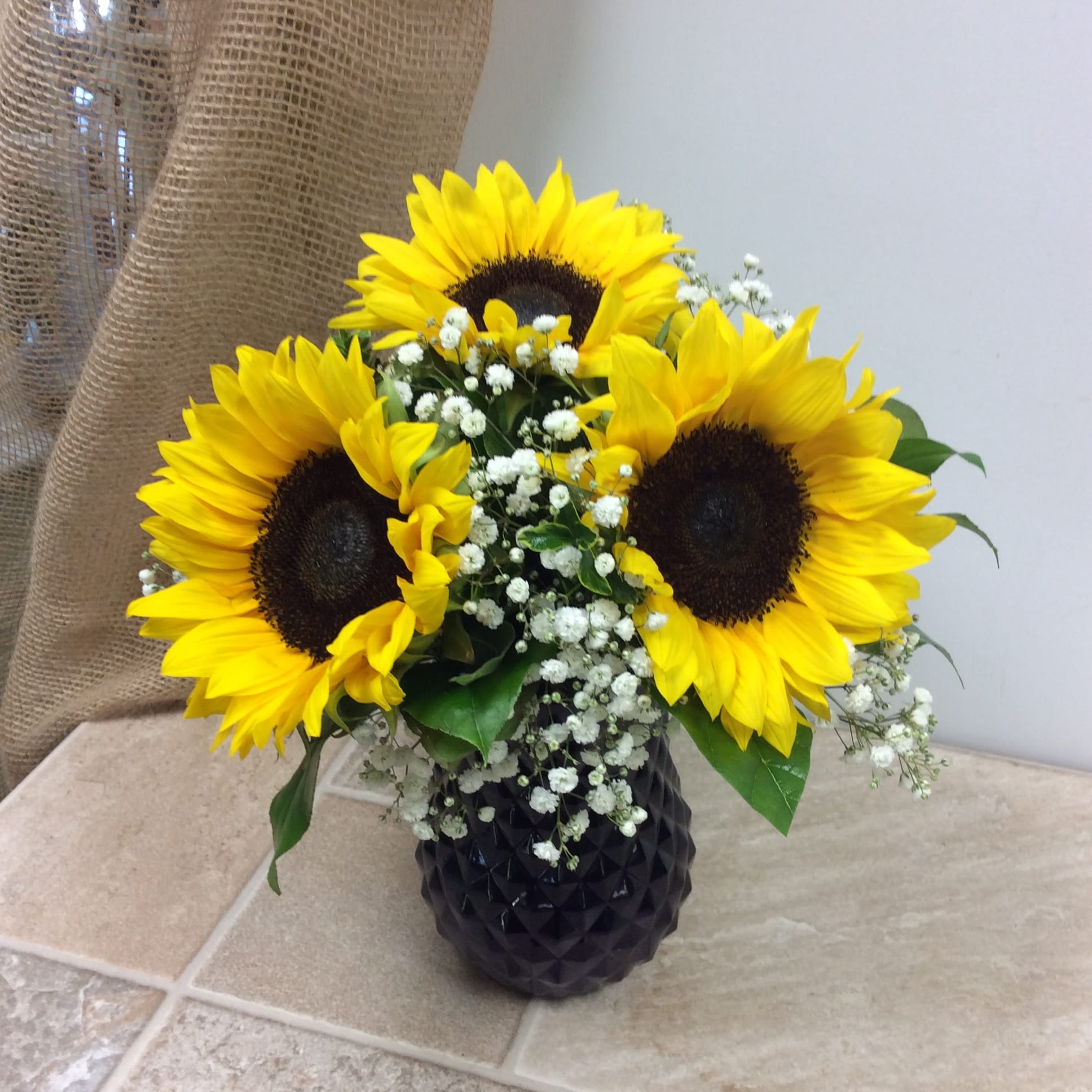 Felix - This happy little bouquet is highlighted by three large sunflowers sure to bring a smile to anyone's face.  The bright blue vase and and baby’s breath complete the summery arrangement.  Primary flowers: sunflower
