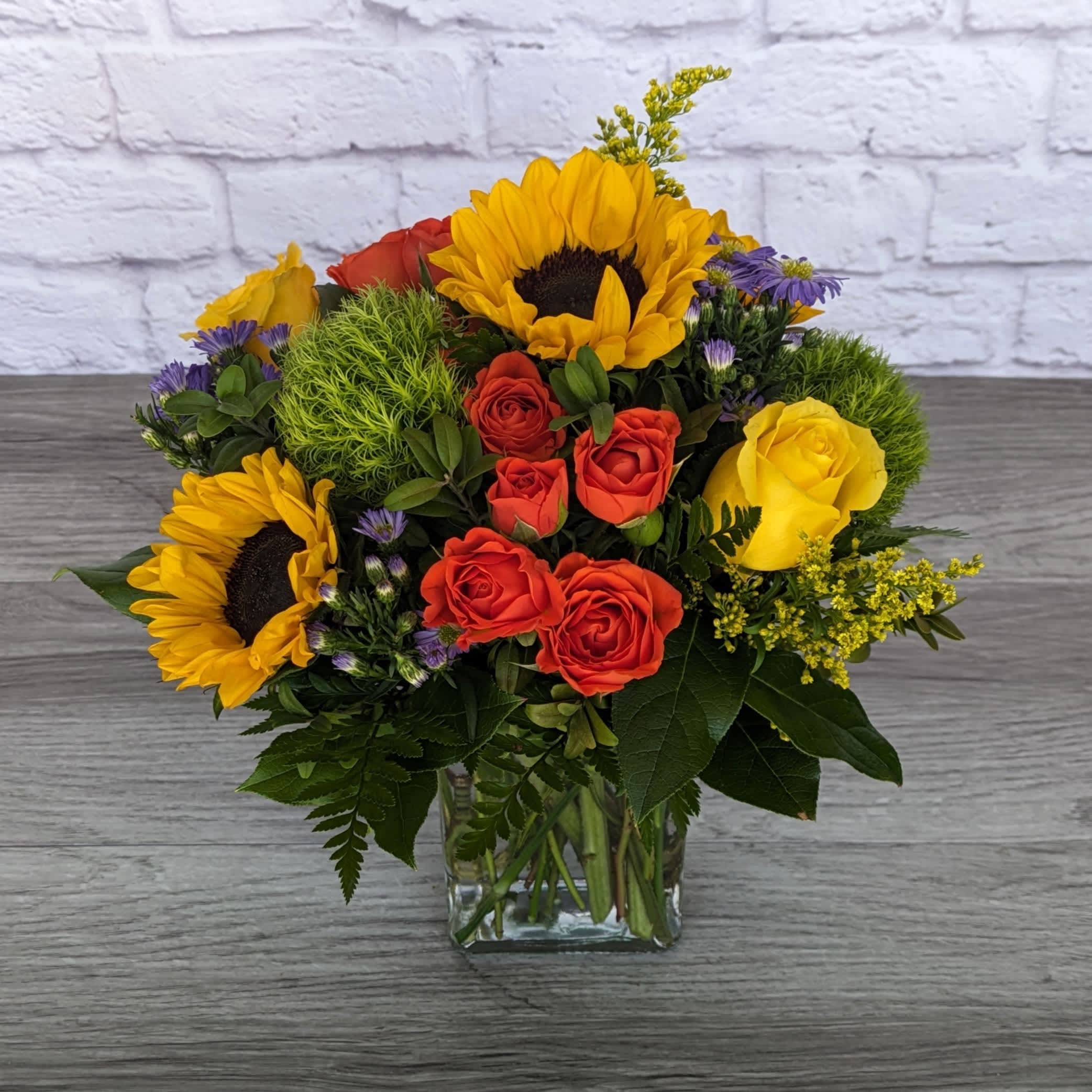 Bonfire Nights - Bonfire Nights is A Ladybug Floral exclusive bouquet inspired by sunsets around the campfire. Featuring Sunflowers, roses, spray roses, green dianthus, monte casino aster and solidago. All within a 6” tall clear rectangular cube.  One Sided Orientation 14"H X 12"W 