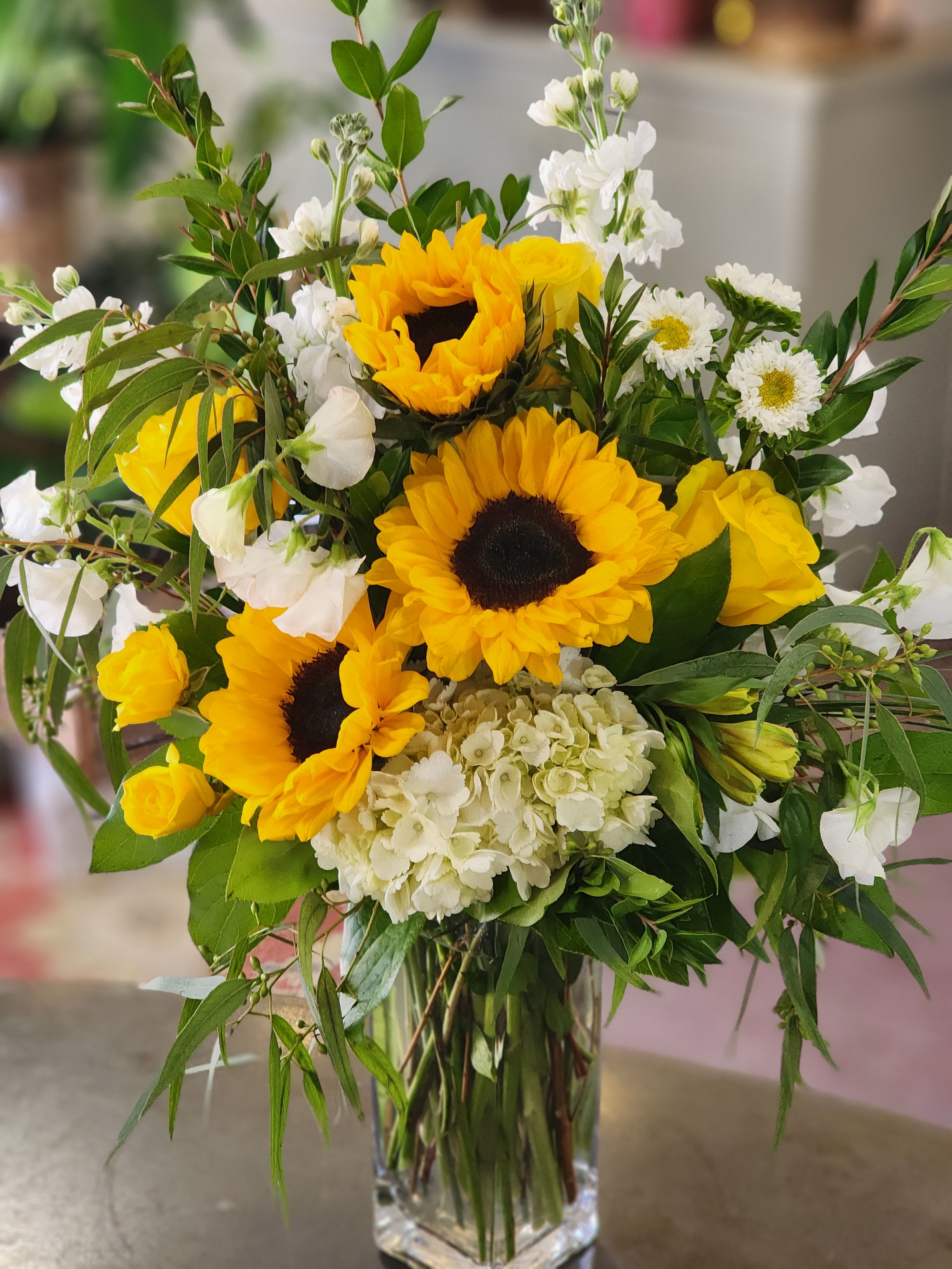 Just to see you smile - This tall square glass vase filled with sunshine is sure to make that special someone smile. Beautiful sunflowers,  hydrangeas, roses, alstromeria, stock and spray roses and wispy greenery create this bouquet of sunshine.