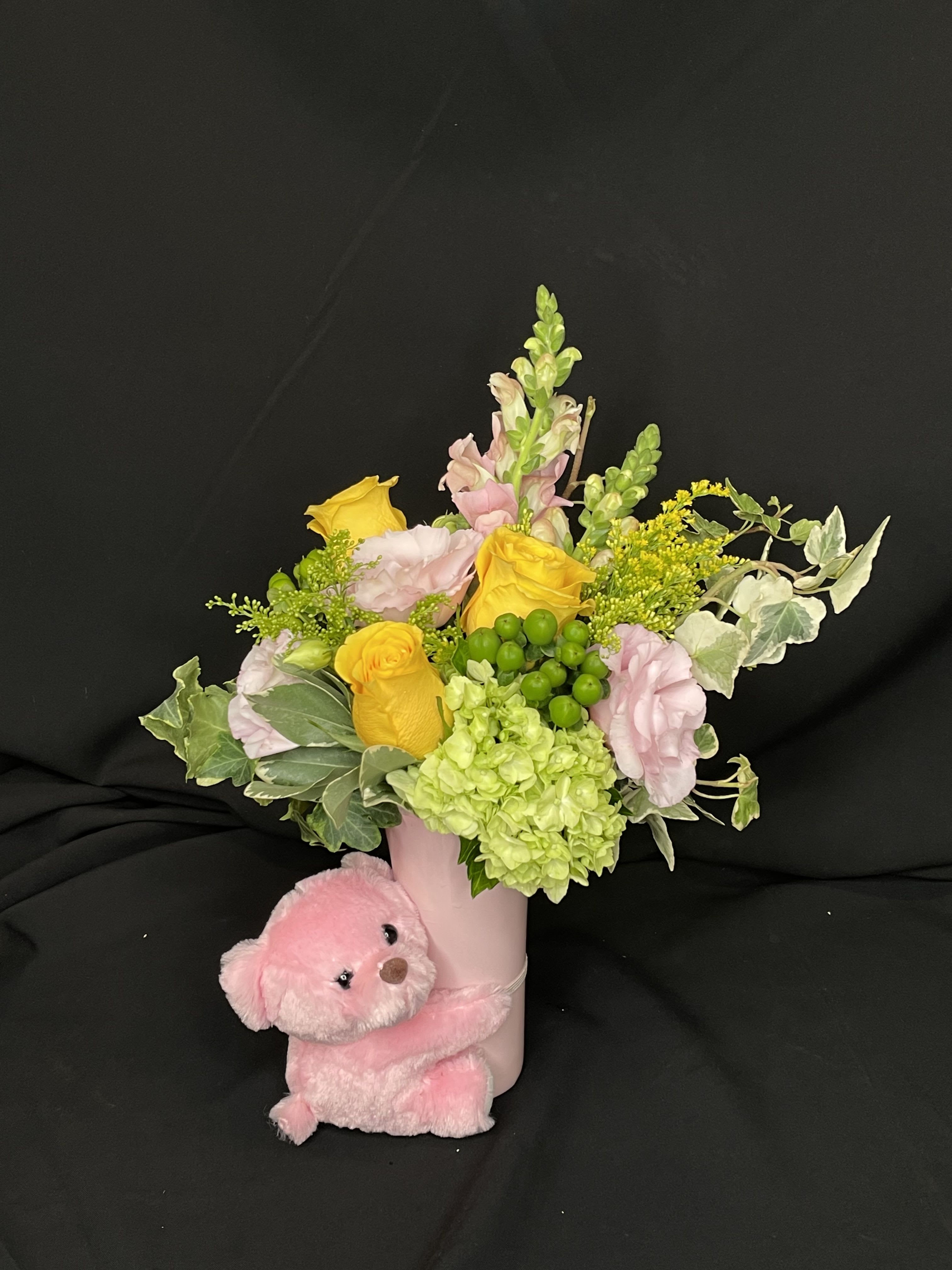 Cuddle Up - A refreshing fusion of sunshine and sweetness! This charming arrangement features cheerful yellows, pinks and greens. The flowers in this arrangement include, roses, lisianthus, snapdragons, hydrangea, solid aster, hypericum berries, and ivy. An adorable pink plush bear adds an extra dose of sweetness to this already delightful design. Perfect for baby showers, sympathy, or brightening someone’s day with fresh, uplifting beauty.  