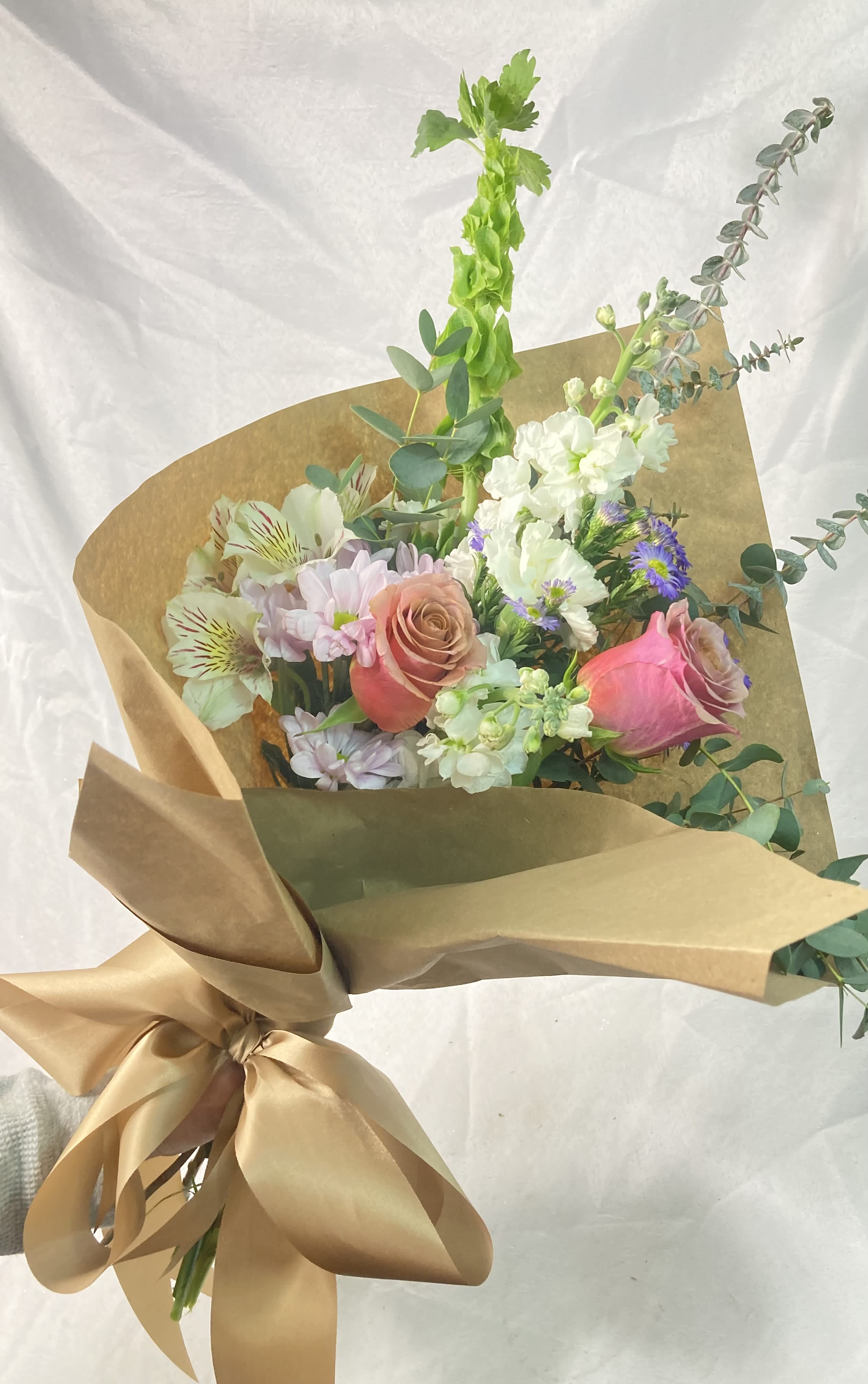 Farm Girl Bouquet - “Farm Girl Bouquet ” is a hand-tied, wrapped bouquet featuring Purple Moon Roses, Stock,  Daisies, Eucalyptus varieties, Chrysanthemums, Alstroemeria, Bells of Ireland &amp; Asters. In a brown paper wrap tied up with a romantic, soft, satin bow, this bouquet will melt her heart! 