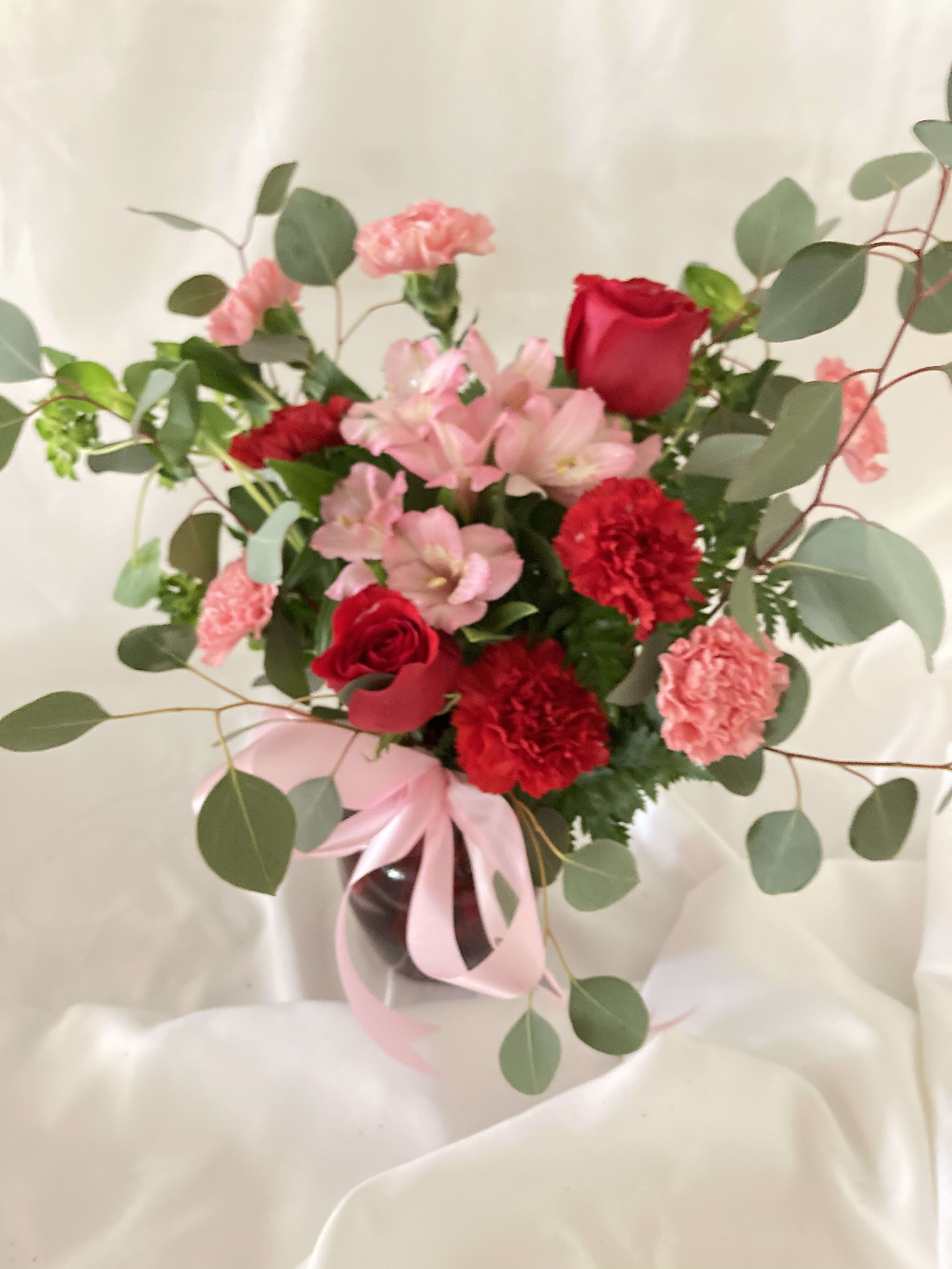 Blooming Belles Bouquet - The belle of the ball! Arranged in a ruby red vase this romantic bouquet of rich red roses and delicate pink alstroemeria is a beautiful statement of love and devotion. Red roses pink alstroemeria red carnations and miniature pink carnations are accented with silver dollar eucalyptus and pitta negra. Delivered in a ruby rose vase.