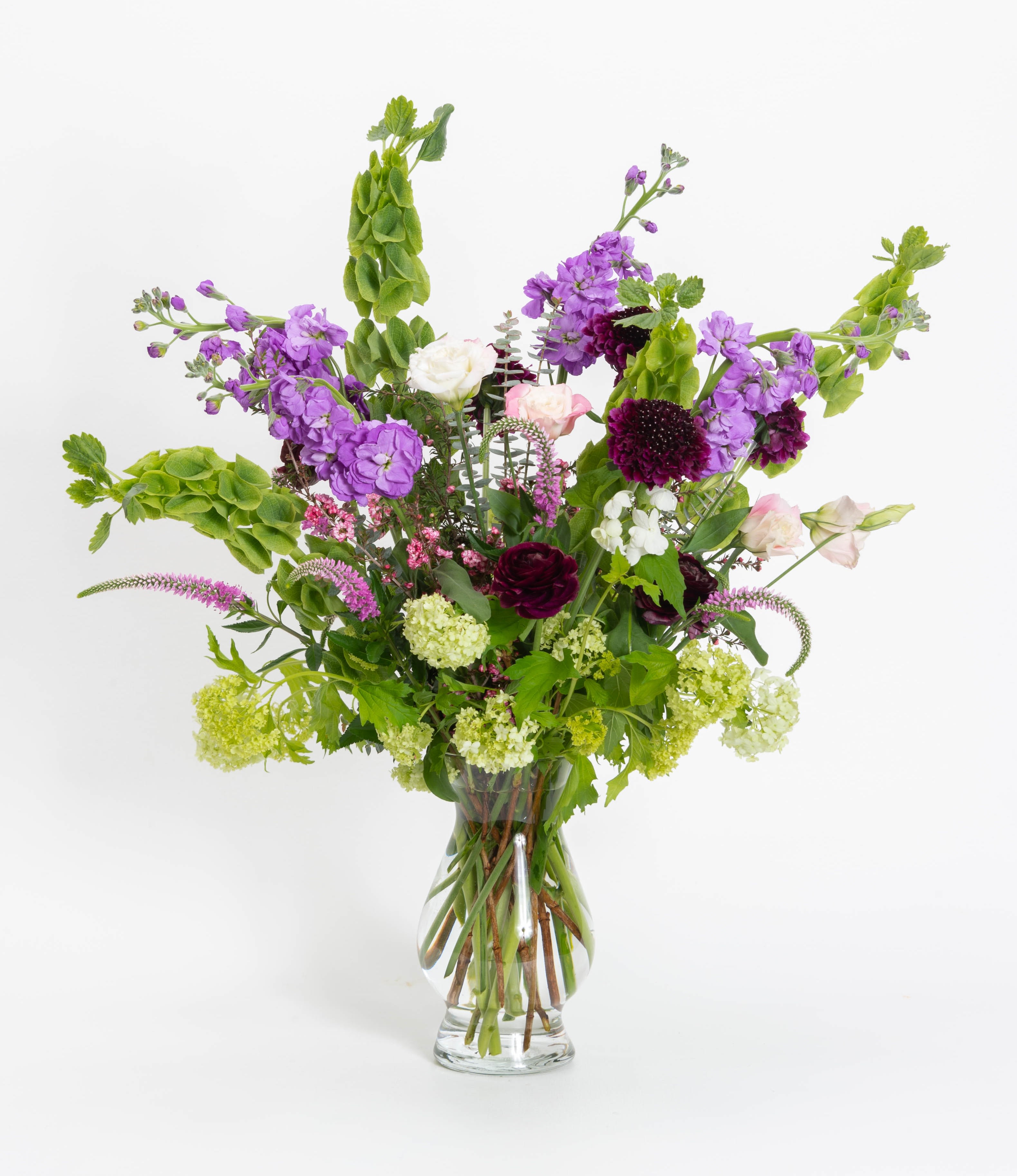 Garden's Path - This tall vase arrangement in purples, lavenders and greens make up this beautiful garden look. Stock, viburnum, bells of Ireland, lisianthis, scabiosa, veronica, ranunculus and lepto. 