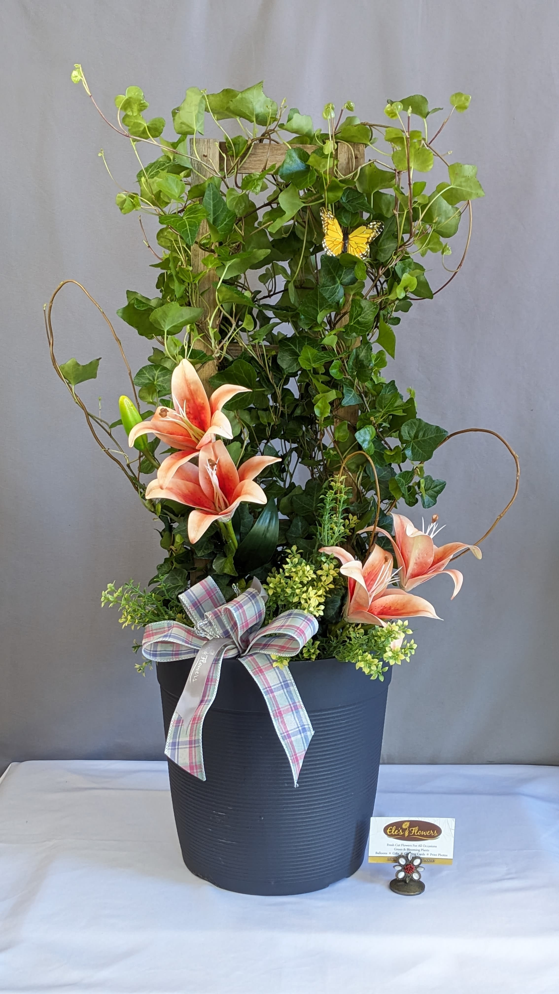 Ivy Embrace - 8" Planter  - Bring height, texture, and lasting beauty into any space with this charming ivy trellis design. Lush, vibrant green ivy climbs naturally up a rustic wooden trellis, creating a graceful vertical display full of movement and life. The rich foliage weaves effortlessly through the frame, giving it a garden-inspired feel that’s both classic and refreshing.  Nestled among the greenery are stunning permanent lily blooms in soft peach and coral tones, adding a warm, elegant pop of color. Delicate accent florals and textured greenery enhance the design, creating depth and visual interest from every angle. A tasteful ribbon detail at the base adds a soft finishing touch, while the modern planter keeps the overall look polished and versatile.  Perfect for home décor, sympathy gifts, offices, or long-lasting seasonal displays, this arrangement offers the beauty of fresh florals with the ease of permanent blooms — a timeless piece that stays vibrant all year long.  Approximate Dimensions: 33"H x 19"W