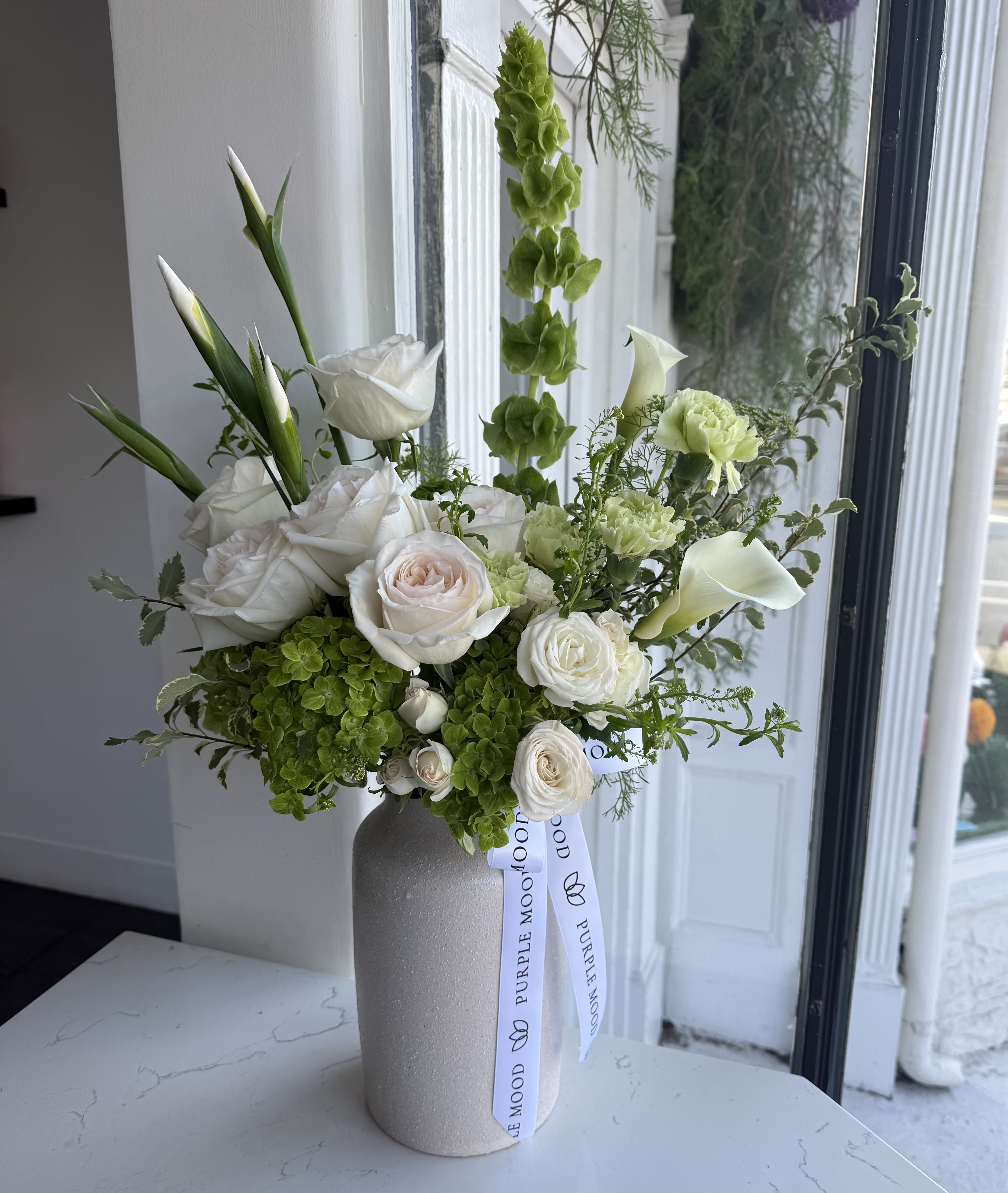 White Bloom Vase - A refined spring arrangement featuring soft ivory roses, fresh green hydrangea, elegant calla lilies, and lush seasonal greenery. Designed in a modern textured vase, this piece captures the beauty of the season with its airy feel and timeless palette.  Perfect for home décor, thoughtful gifting, or celebrating a special moment with effortless elegance.