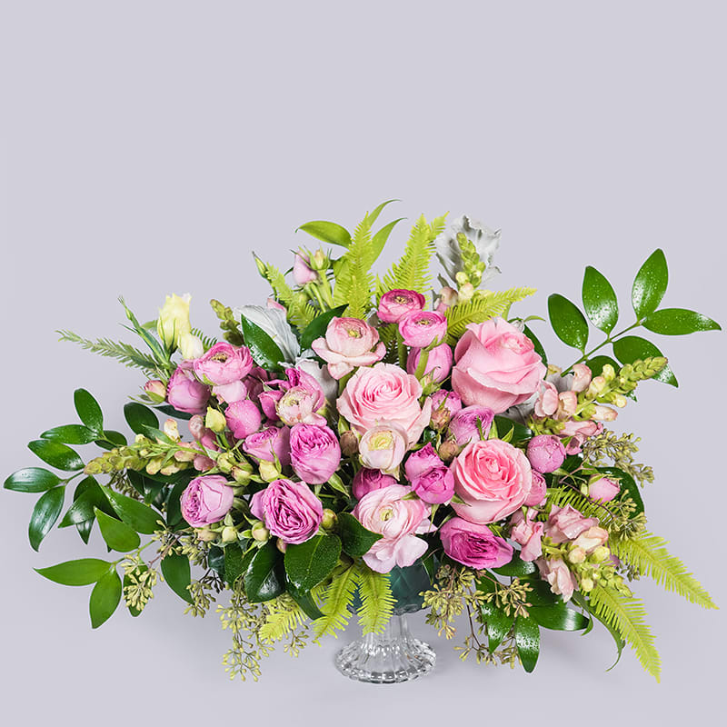 Low pink rose and ranunculus arrangement with snapdragons and bright green ferns in a clear glass vase