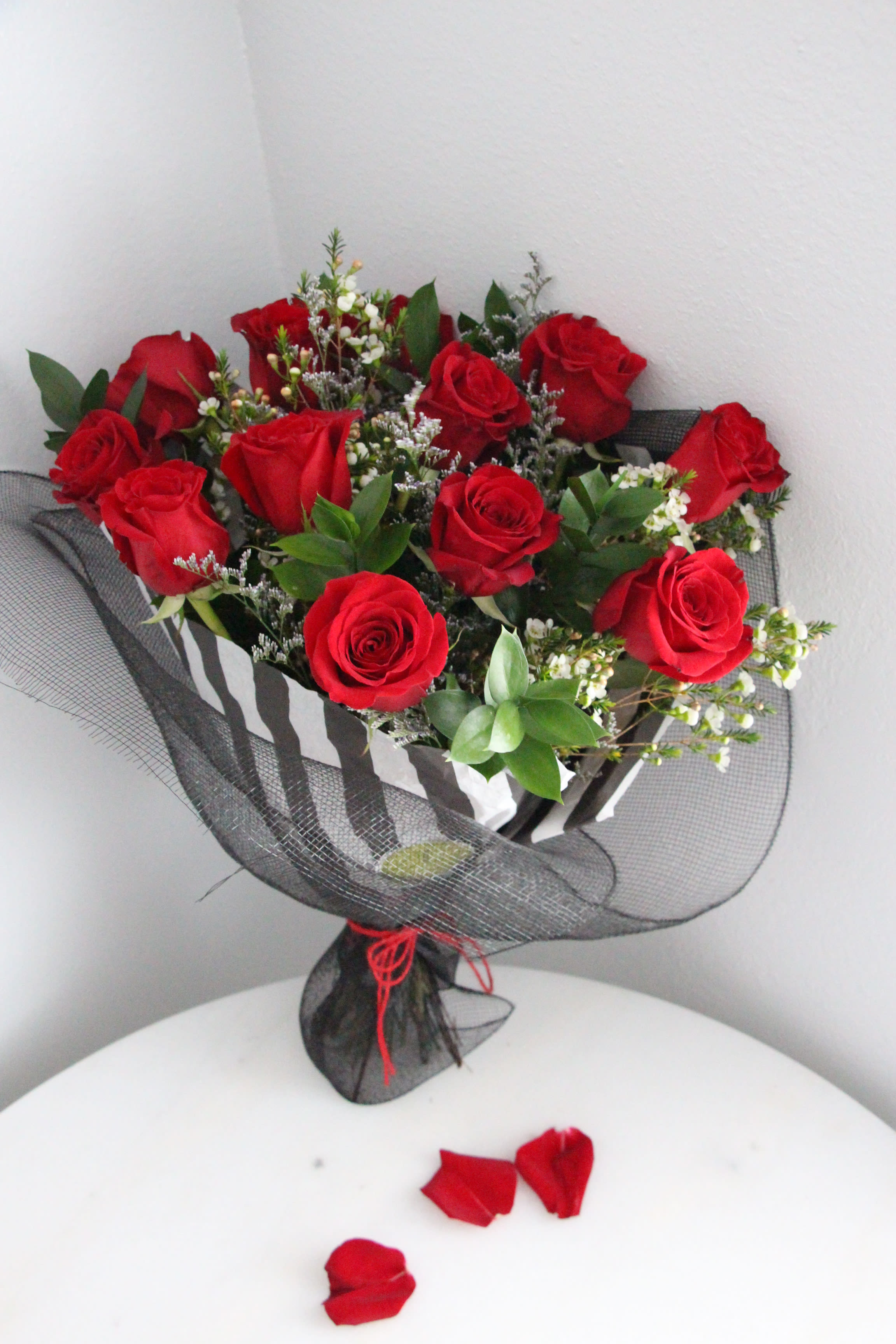 Hand-tied bouquet of red roses with white filler flowers wrapped in black mesh, with rose petals on a table