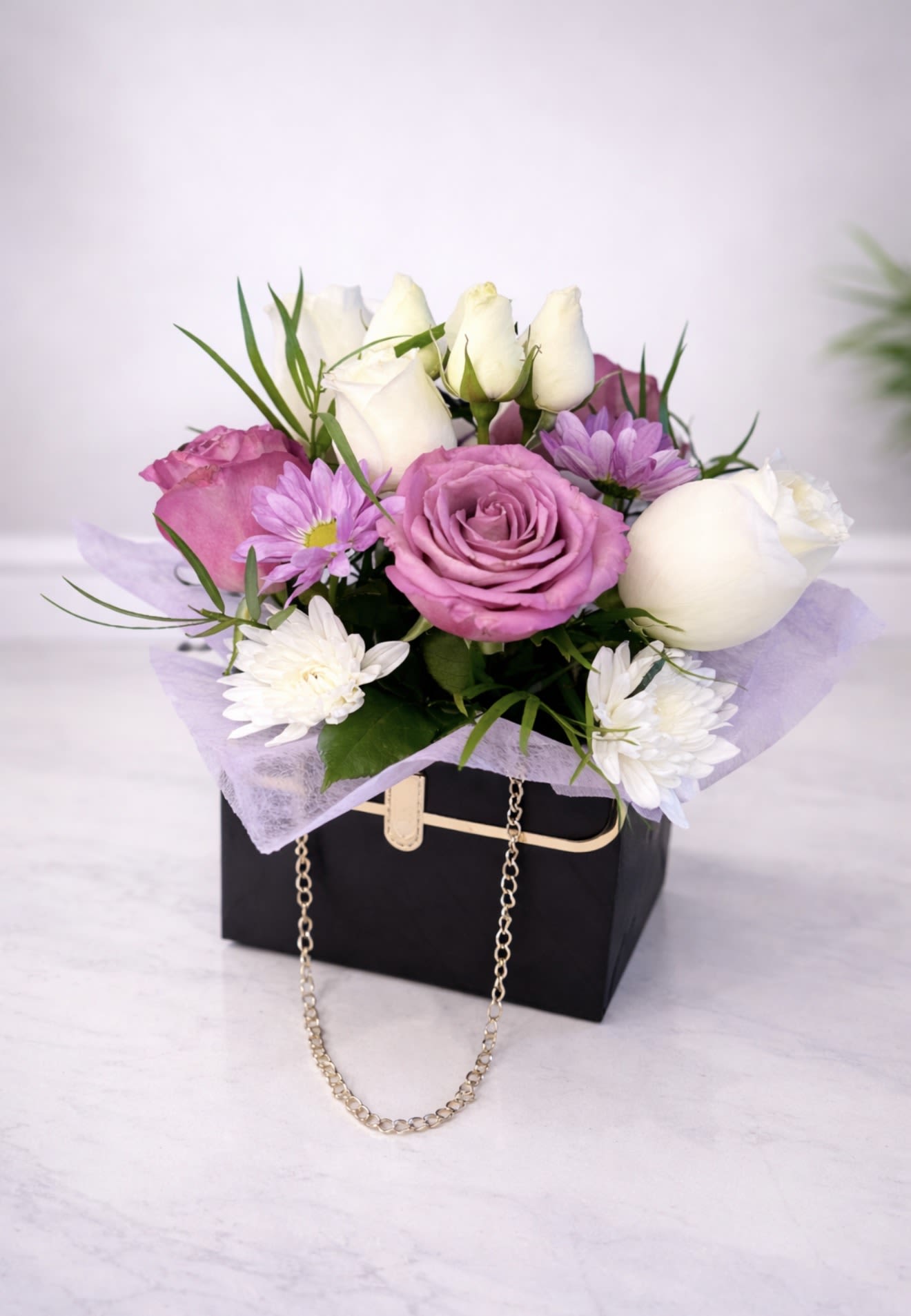 Lavender Luxe Handbag Arrangement - A chic and feminine floral handbag arrangement featuring lavender roses, white roses, soft purple blooms, and fresh greenery designed in an elegant black keepsake box with gold chain detail. This modern, stylish flower arrangement is perfect for birthdays, anniversaries, thank-you gifts, or a luxurious surprise.  Handcrafted with premium fresh flowers and available for same-day flower delivery in Alpharetta, GA 30022.
