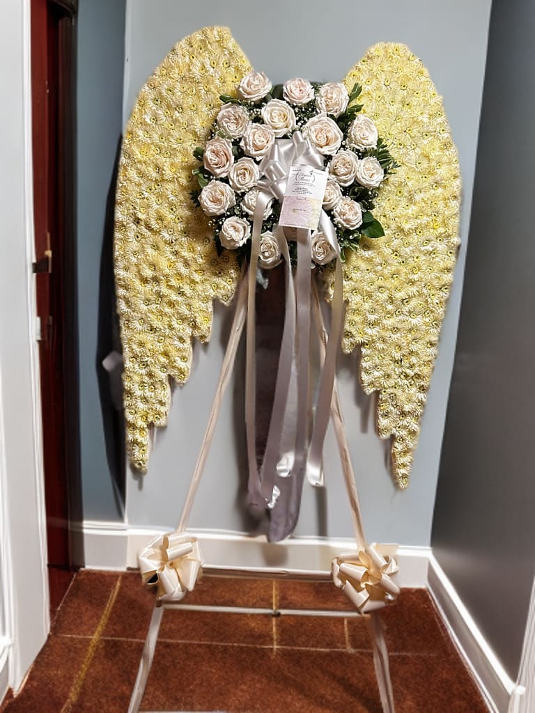 Angel wings white  - This elegant funeral arrangement is designed in the shape of angel wings, symbolizing peace, protection, and eternal rest. The wings are crafted from delicate white and pale yellow flowers, radiating purity and light. At the center, a round cluster of soft ivory roses accented with lush greenery represents love and remembrance. Finished with satin ribbons and bows, this heavenly tribute offers comfort and honors the memory of a loved one with grace and serenity