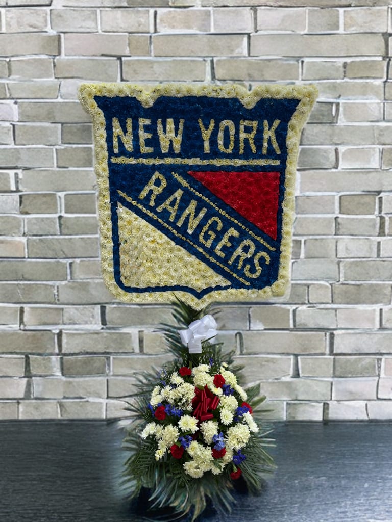 New York Rangers  - This custom funeral arrangement is a heartfelt tribute for a devoted New York Rangers fan. Designed in the shape of the team’s iconic logo, it is carefully crafted with fresh flowers in the team colors of blue, red, and white. At the base, a lush spray of red, white, and blue blooms accented with greenery and a white bow adds elegance. A meaningful way to honor a loved one’s passion for their favorite team, it reflects both pride and remembrance in a beautiful, dignified display.