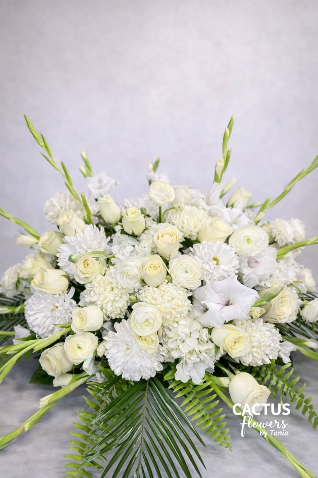 Heavenly Light Tribute - A graceful all-white sympathy arrangement designed with white roses, ranunculus, chrysanthemums, gladiolus and fresh greenery. A timeless expression of peace, purity and remembrance.