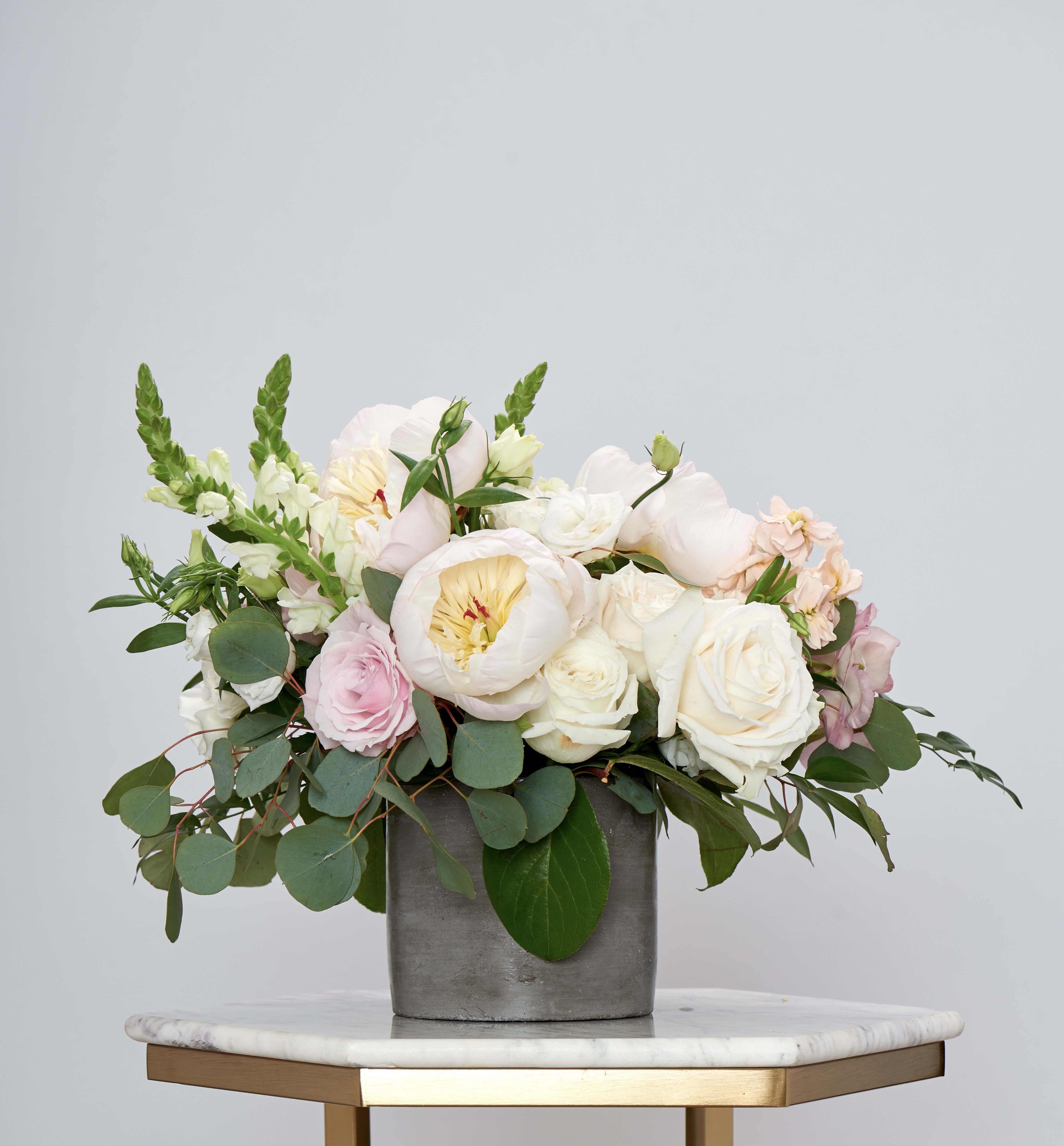 Peony Pier - Peony Pier is a refined garden-style arrangement designed in soft blush and ivory tones with seasonal blooms and lush greenery. Set in a modern vessel, this design blends romance with understated elegance, creating a look that feels both timeless and elevated.  Perfect for anniversaries, milestone birthdays, bridal celebrations, or meaningful romantic gestures, Peony Pier offers a graceful way to send something beautiful and memorable.  Designed using the freshest seasonal flowers available. While flower varieties may vary slightly, the overall palette, size, and garden-inspired style will remain true to the photo shown.  Photo displays deluxe size.