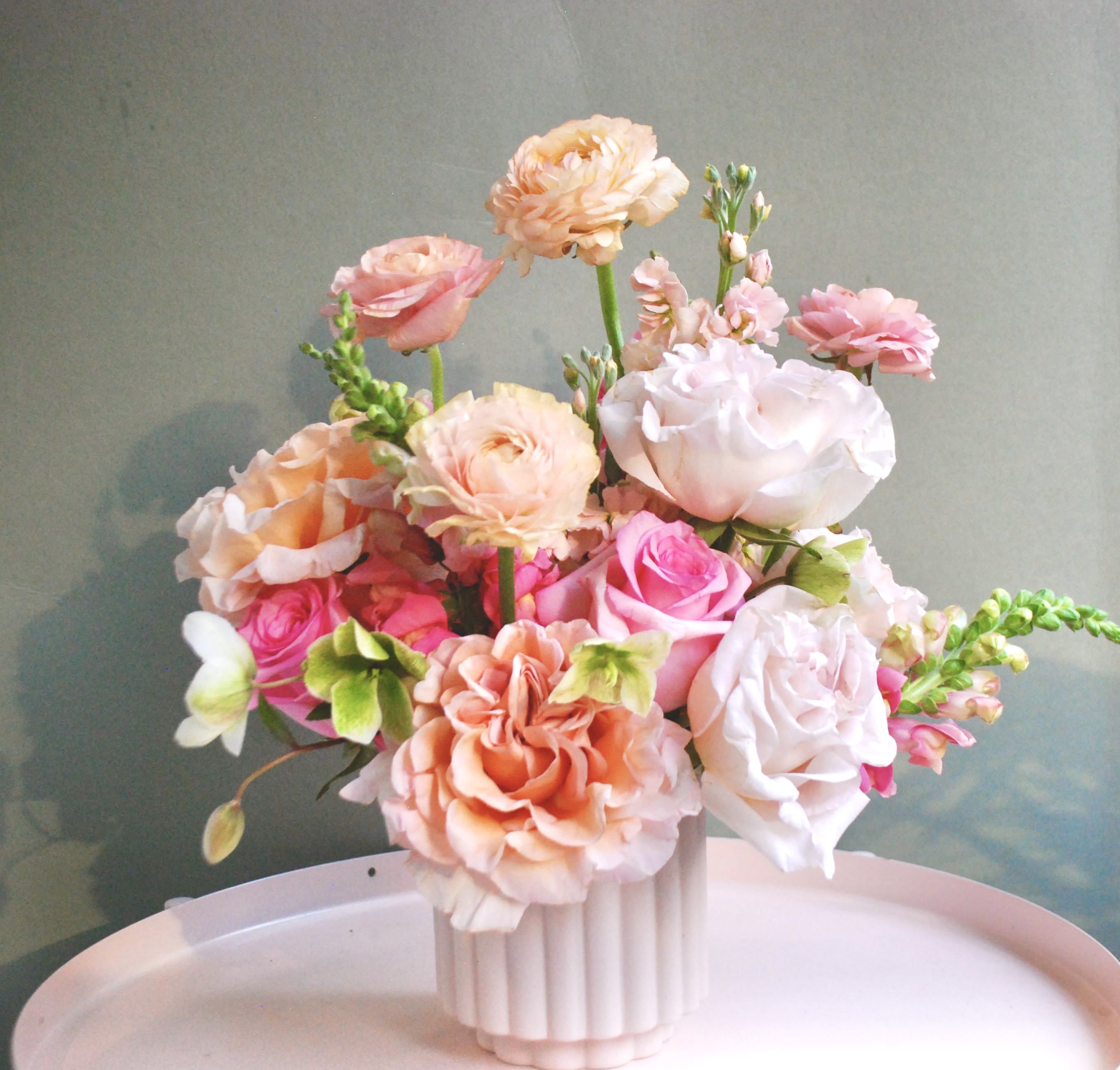 Dive -  A soft, romantic arrangement designed in a modern white ribbed ceramic vase. This piece features a delicate mix of blush and peach garden-style blooms, including ranunculus, roses, lisianthus, and seasonal accent flowers. Airy textures and gentle movement give this arrangement a light, elegant feel, perfect for brightening a space without overwhelming it.