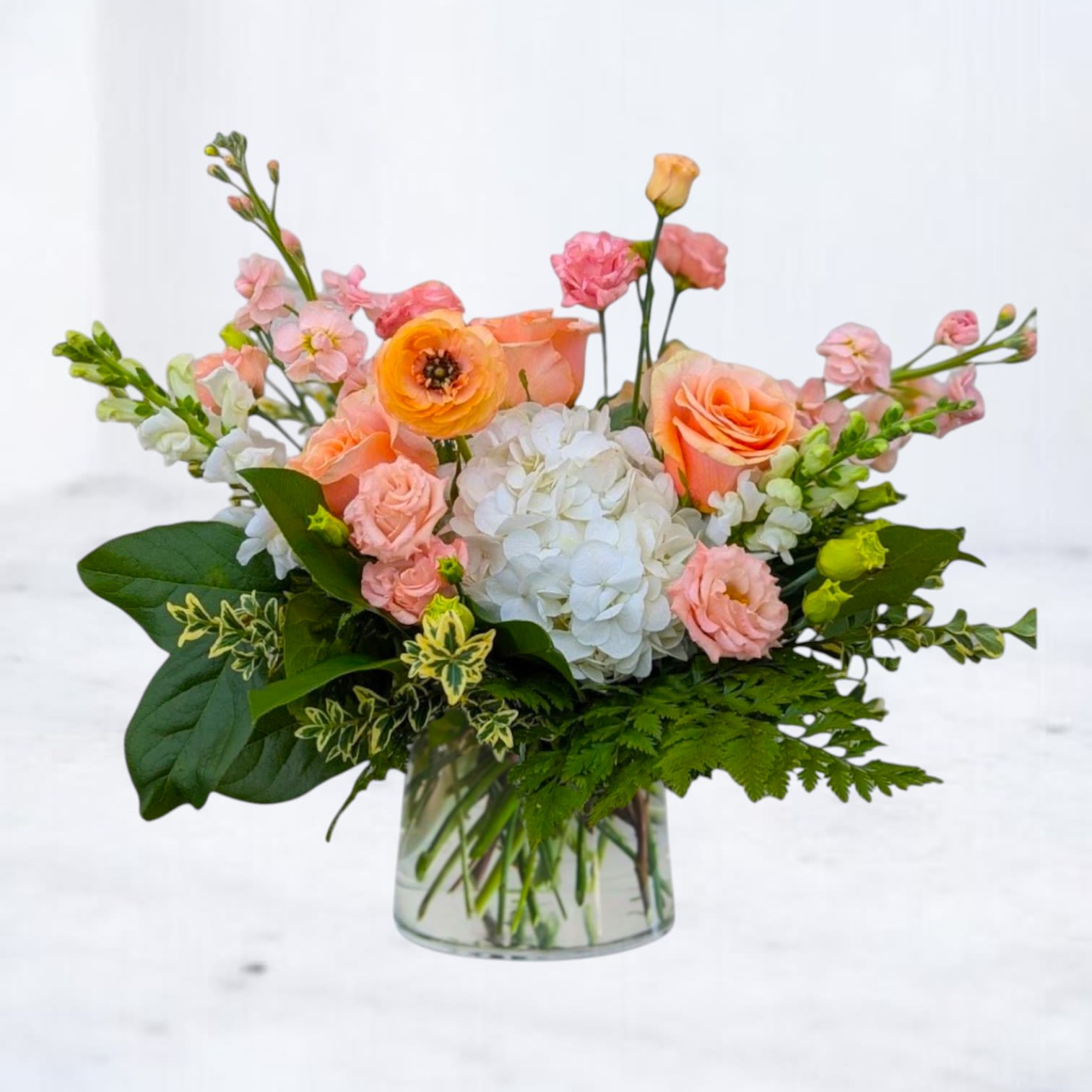 North Apricot - North Apricot is a refined garden-style arrangement featuring peach roses, blush spray roses, white hydrangea, and seasonal blooms layered with fresh greenery for natural movement and texture. The soft apricot tones are balanced with crisp white florals, creating a design that feels warm, modern, and effortlessly composed.  Designed in a clear glass vase, North Apricot is perfect for birthdays, thank-you gifts, new baby celebrations, or sending something beautiful just because. Its soft peach palette and organic structure make it a versatile choice for both home and office settings.  Available for same-day flower delivery in Magnolia, The Woodlands, Spring, Conroe, and throughout the Greater Houston area. Flower varieties may vary based on seasonal availability while maintaining the overall color story and fullness of the design.