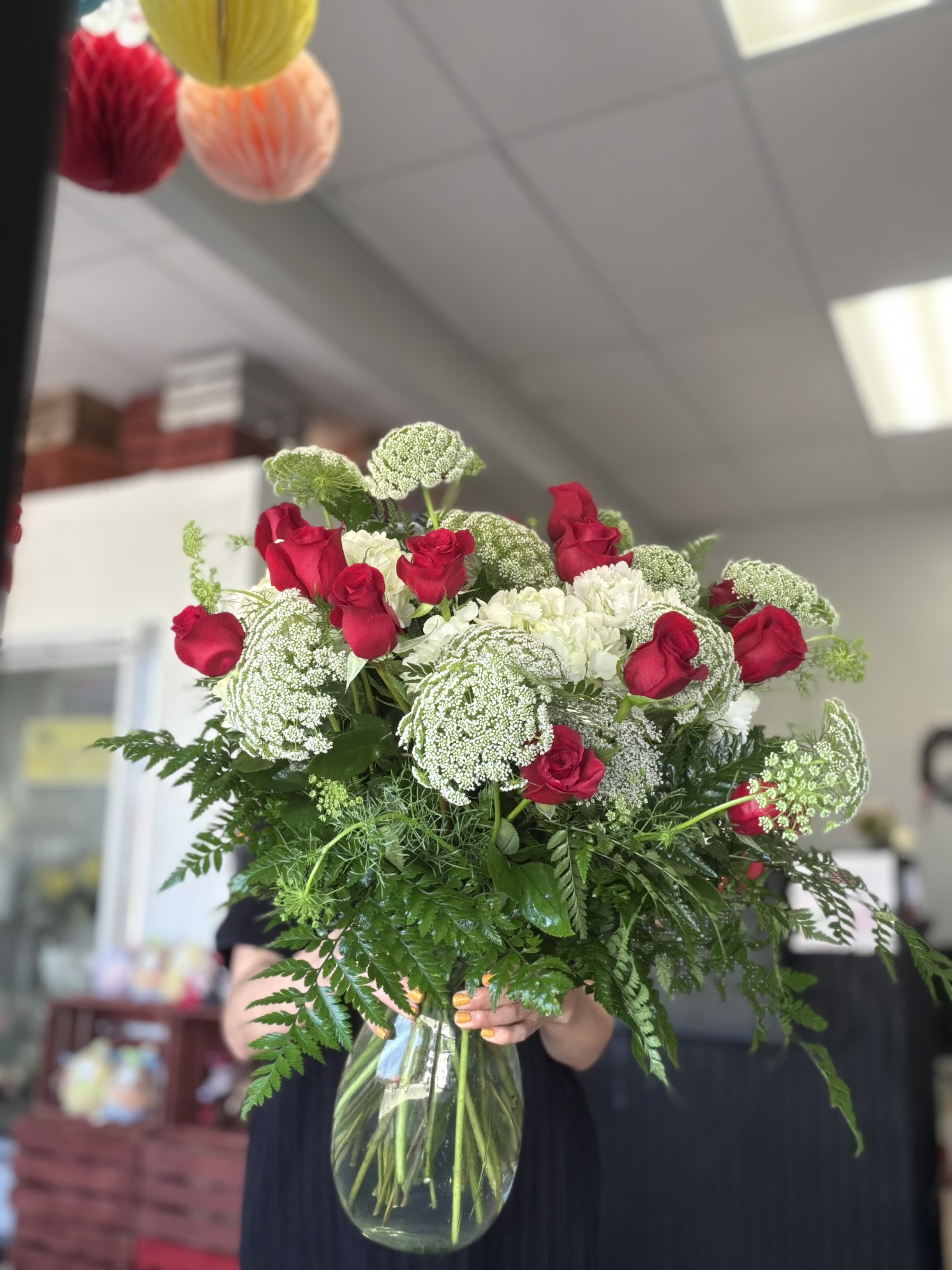 Crimson Lace Reverie - Make a bold yet timeless statement with this lush, hand-crafted arrangement featuring classic red roses beautifully accented with delicate Queen Anne’s lace and crisp white blooms. Designed with abundant fresh greenery for a naturally full and airy look, this bouquet blends romance with organic texture for a refined, garden-inspired feel.  Arranged in a clear glass vase, the vibrant red roses create a striking contrast against the soft white floral queen anne lace, making it perfect for anniversaries, romantic gestures, birthdays, or simply elevating any space with fresh elegance.  Each arrangement is thoughtfully designed to feel generous, balanced, and effortlessly sophisticated — a stunning centerpiece that leaves a lasting impression. 