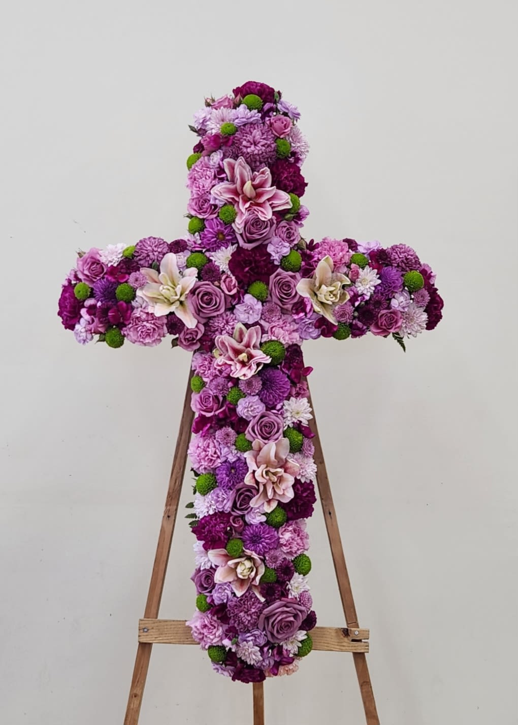 Thoughtful Reflection Cross - Celebrate a life well-lived with this stunning memorial cross, crafted from an elegant blend of purple blooms. Featuring graceful lilies, full-bodied carnations, classic roses, delicate spray roses, and cheerful mums, each flower is thoughtfully arranged to create a meaningful tribute. The rich purple palette conveys dignity and remembrance, making this piece a compassionate choice for services, funerals, or to honor someone special. A beautiful way to express your deepest condolences and support during difficult times.
