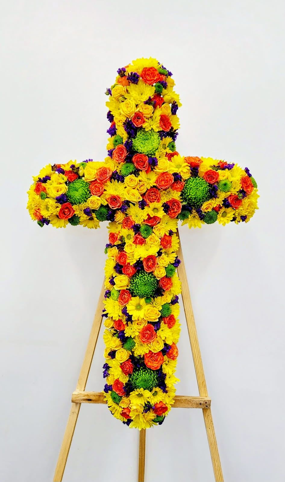 Shine Brightly Cross - Celebrate a life well-lived with the Shine Brightly Cross, a radiant tribute arrangement featuring vibrant yellow blooms accented with touches of warm orange and lush green mums. This elegant standing cross combines bold, cheerful blooms with sophisticated design, creating a meaningful memorial that honors cherished memories with grace and warmth. Perfect for services, viewings, or as a heartfelt expression of sympathy.