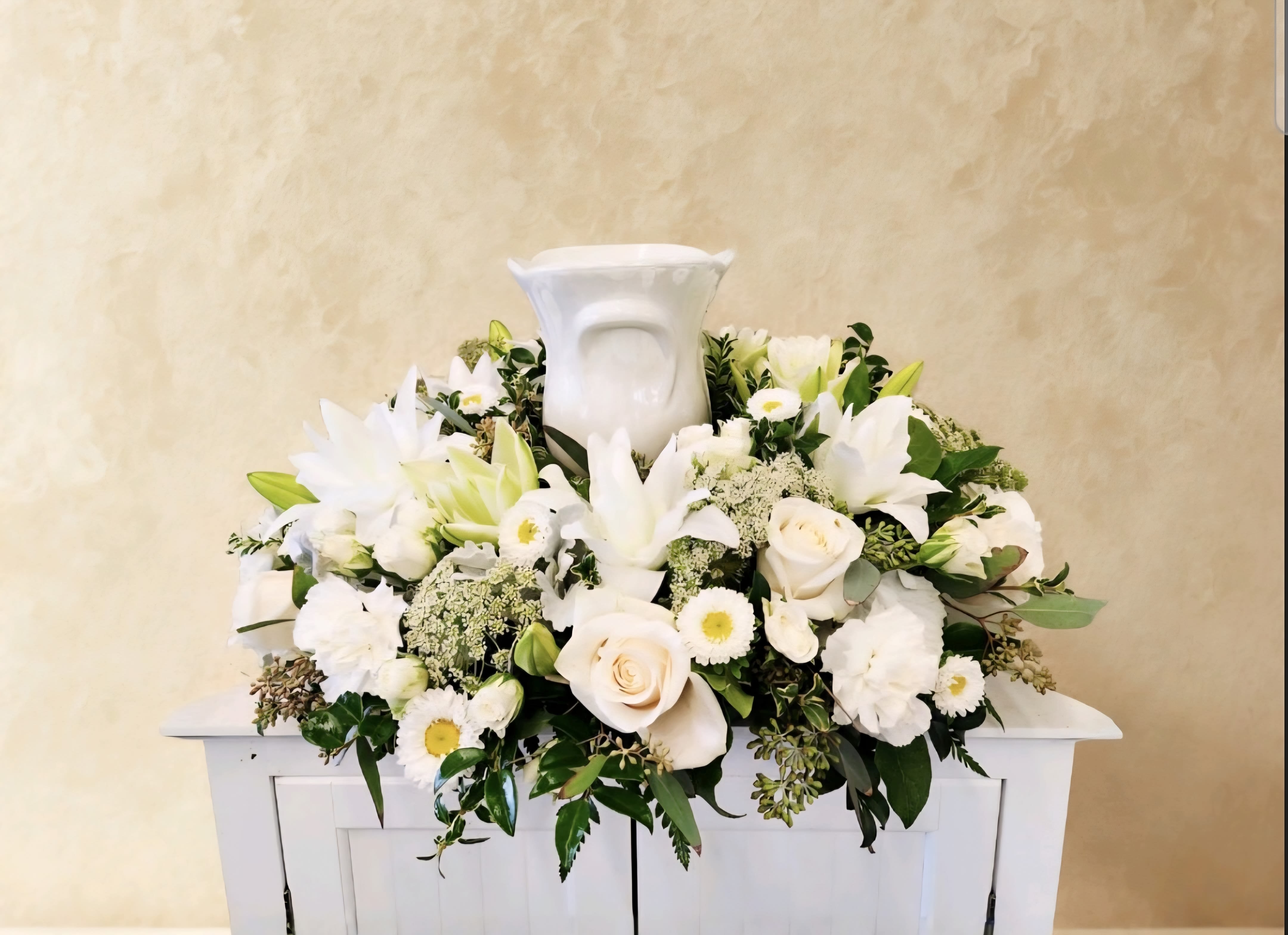 Fond Memories - Fond Memories is a serene all-white cremation arrangement that honors your loved one with timeless elegance. This thoughtfully composed piece features premium roses, lilies, and carnations layered with delicate mums and Queen Anne's lace, all accented by lush greenery. Perfect for memorial services, this arrangement conveys compassion and respect during a meaningful time.  One Size Only: 18" wide by 8" Tall