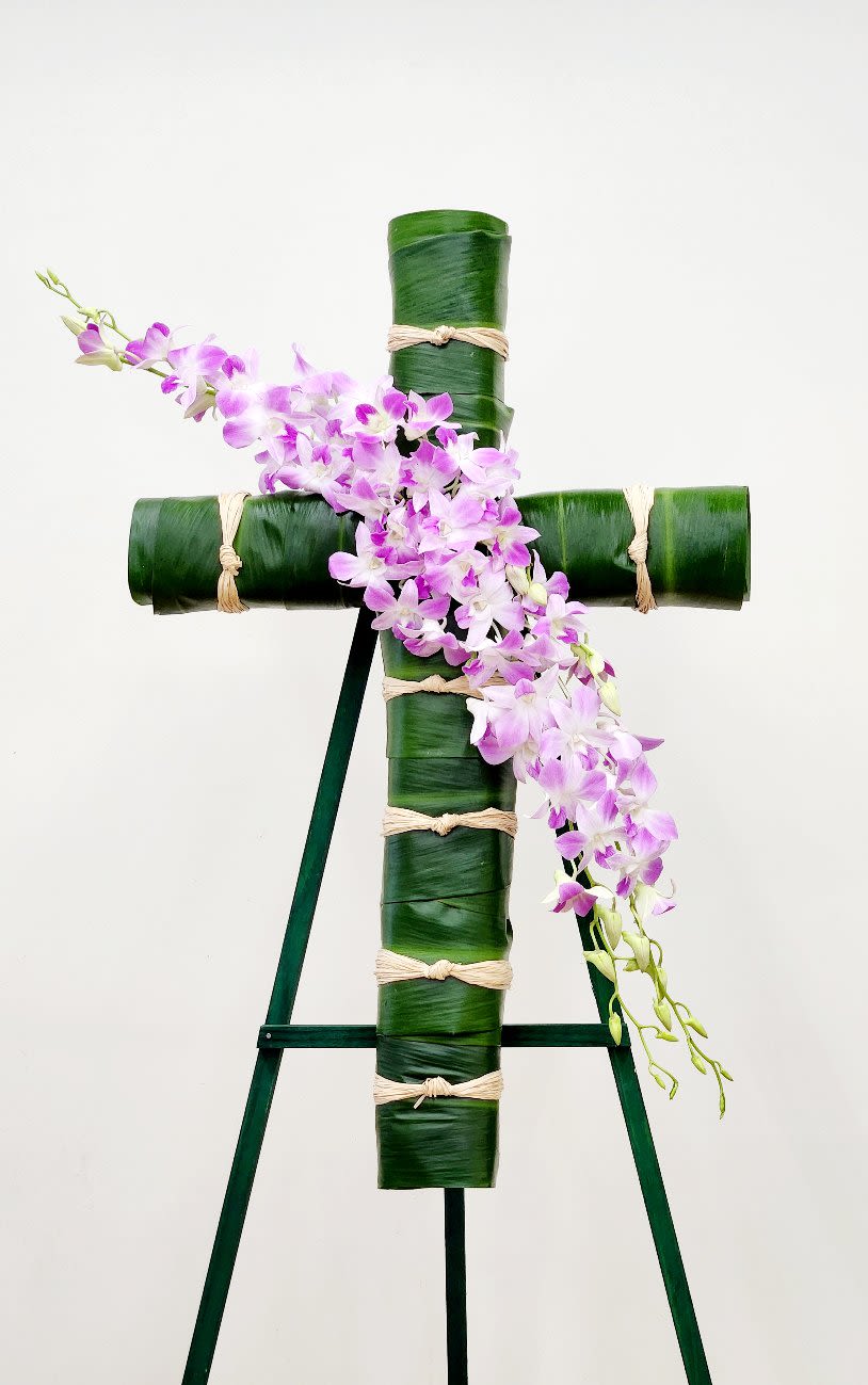 Farewell Cross - A striking tribute arrangement featuring a cross form wrapped in verdant ti leaves, with lavender orchids cascading gracefully across its face. This piece balances natural elegance with understated warmth, creating a distinctive memorial that speaks to both reverence and beauty. Each element is thoughtfully placed to honor and celebrate a life well-lived. ONE SIZE ONLY. 32" CROSS.