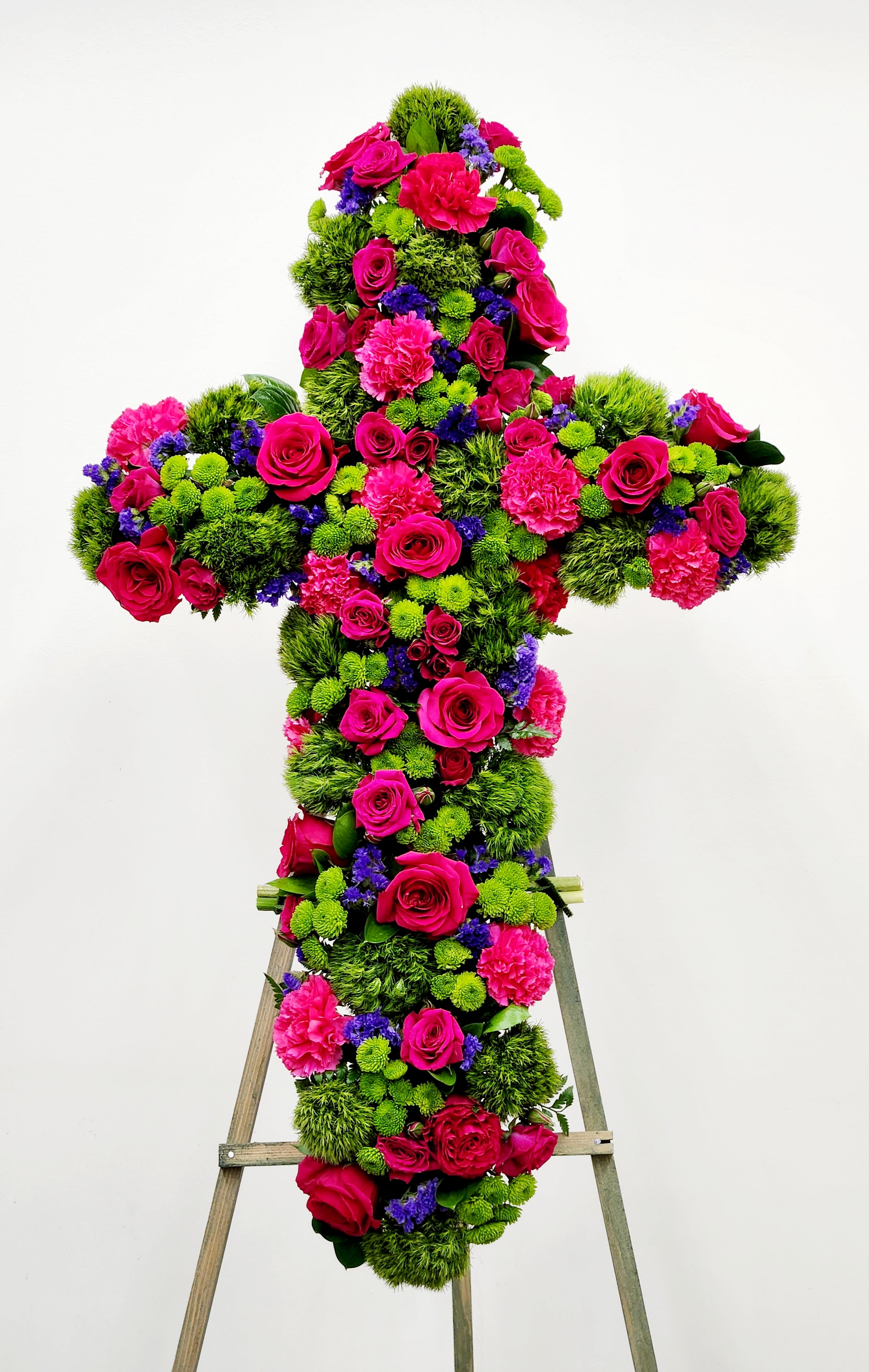 Sweet Goodbye Cross - A timeless floral cross honoring cherished memories with grace and elegance. Lush blooms of green hydrengeas, mums and dainty green buttons form the foundation, complemented by deep pink blooms that convey compassion and respect. A subtle whisper of blue adds depth and serenity to this meaningful tribute. Perfect for expressing sympathy and celebrating a life well-lived.