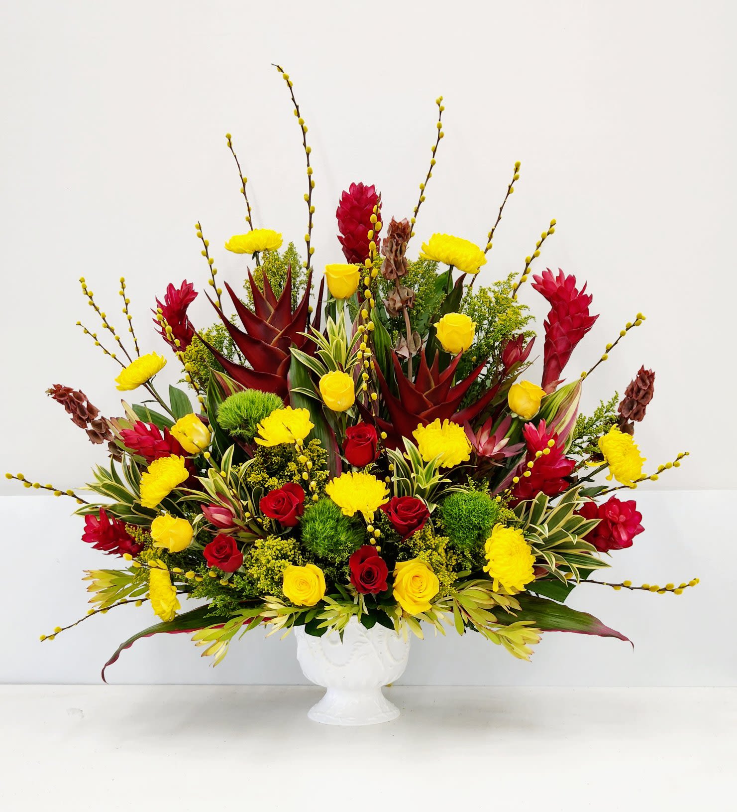 Tropical Elegance - Celebrate their special day with vibrant tropical blooms that radiate warmth and joy. This sophisticated arrangement features striking red ginger and heliconia, complemented by cheerful yellow mums, green dianthus, and golden roses. Lush exotic greenery adds depth and texture, while the elegant white vase provides a refined backdrop for this show-stopping composition. Perfect for making any occasion unforgettable.  Approximate Size:  Standard: 20" x 25"  Deluxe: 25" x 35"  Premium - As Pictured: 32" x 42"