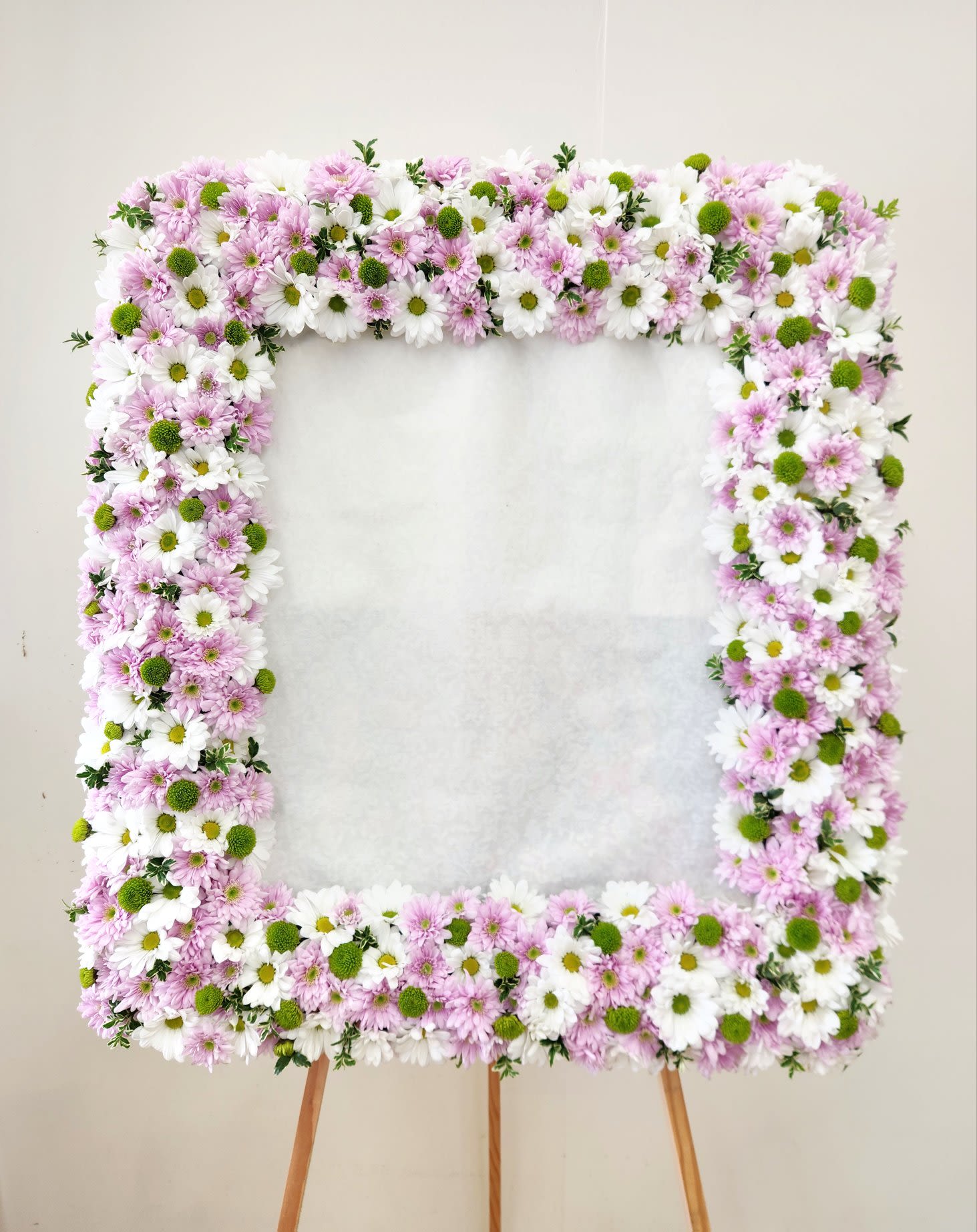 Custom Photo Frame Daisies White and Lavender - A timeless tribute crafted with fresh blooms arranged around a cherished photograph. This custom floral frame transforms a beloved image into a living memorial, perfect for displaying at services, celebrations of life, or intimate gatherings. Each arrangement is thoughtfully designed to honor your loved one with natural beauty and elegance. An meaningful way to keep their memory present and celebrated. ONE SIZE ONLY.  PICTURE SIZE: 24" Tall by 22" Wide  Finished Photo Frame 36" Tall by 30" Wide  FOR SPECIFIC FLOWERS AND COLORS, PLEASE CONTACT THE SHOP AT 562-426-6996  OR VISIT OUR LOCATION AT 611 EAST CARSON STREET, SUITE B, LONG BEACH, CA, 90807