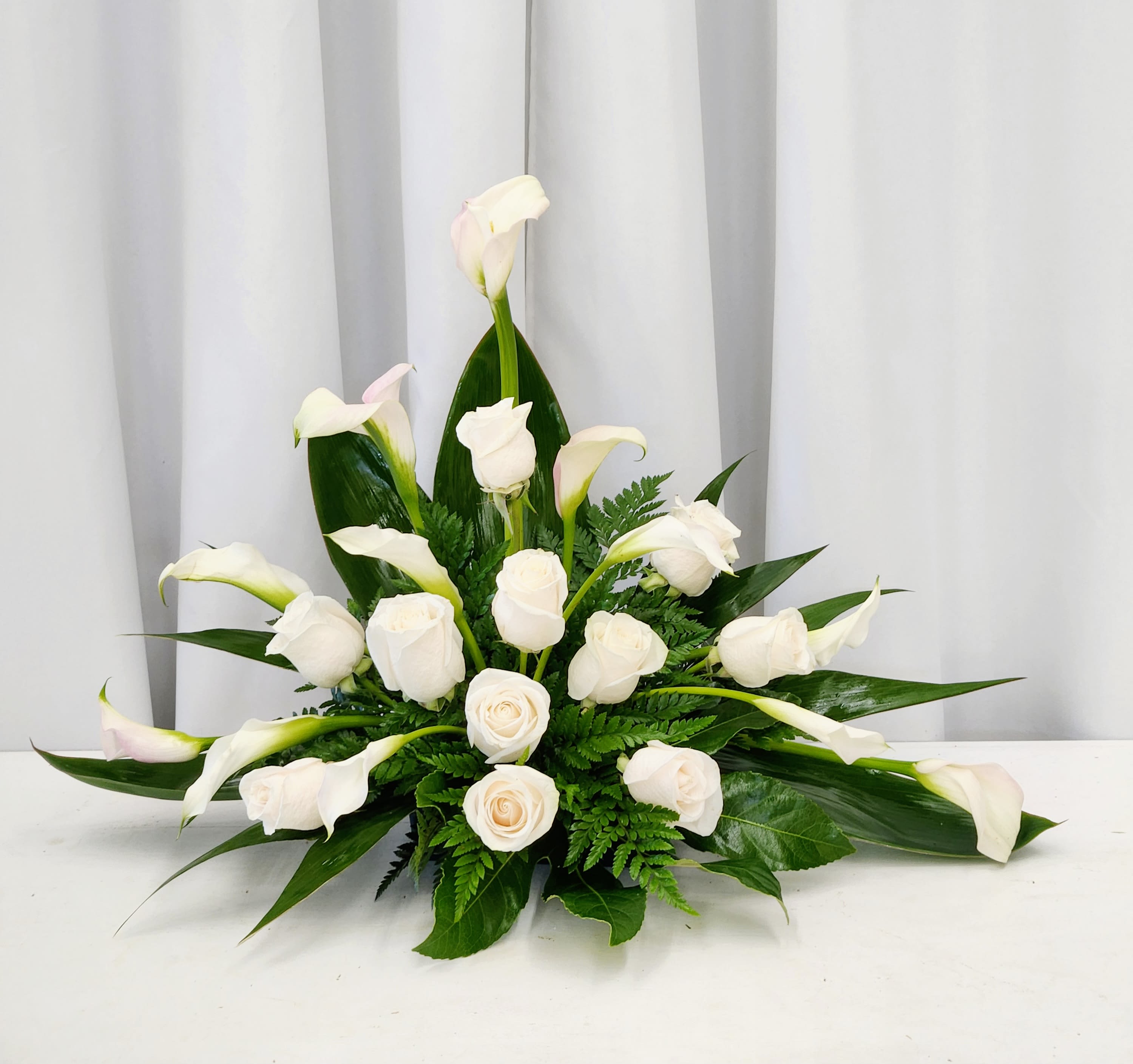 Blossoms of Peace - A serene tribute designed to honor cherished memories and express deepest sympathy. This cremation arrangement features graceful white calla lilies and pristine white roses, softly accented with delicate green ferns and ti leaves. Perfect for placement beside a photograph or urn, it creates a peaceful focal point during times of remembrance. Each bloom is thoughtfully selected to convey compassion and respect.  One Size Only: 18" Wide by 22" Tall    For custom colors please indicate preference in special instructions or visit our shop at  611 E Carson Street, Long Beach, CA 90807  Tel # 562-426-6996