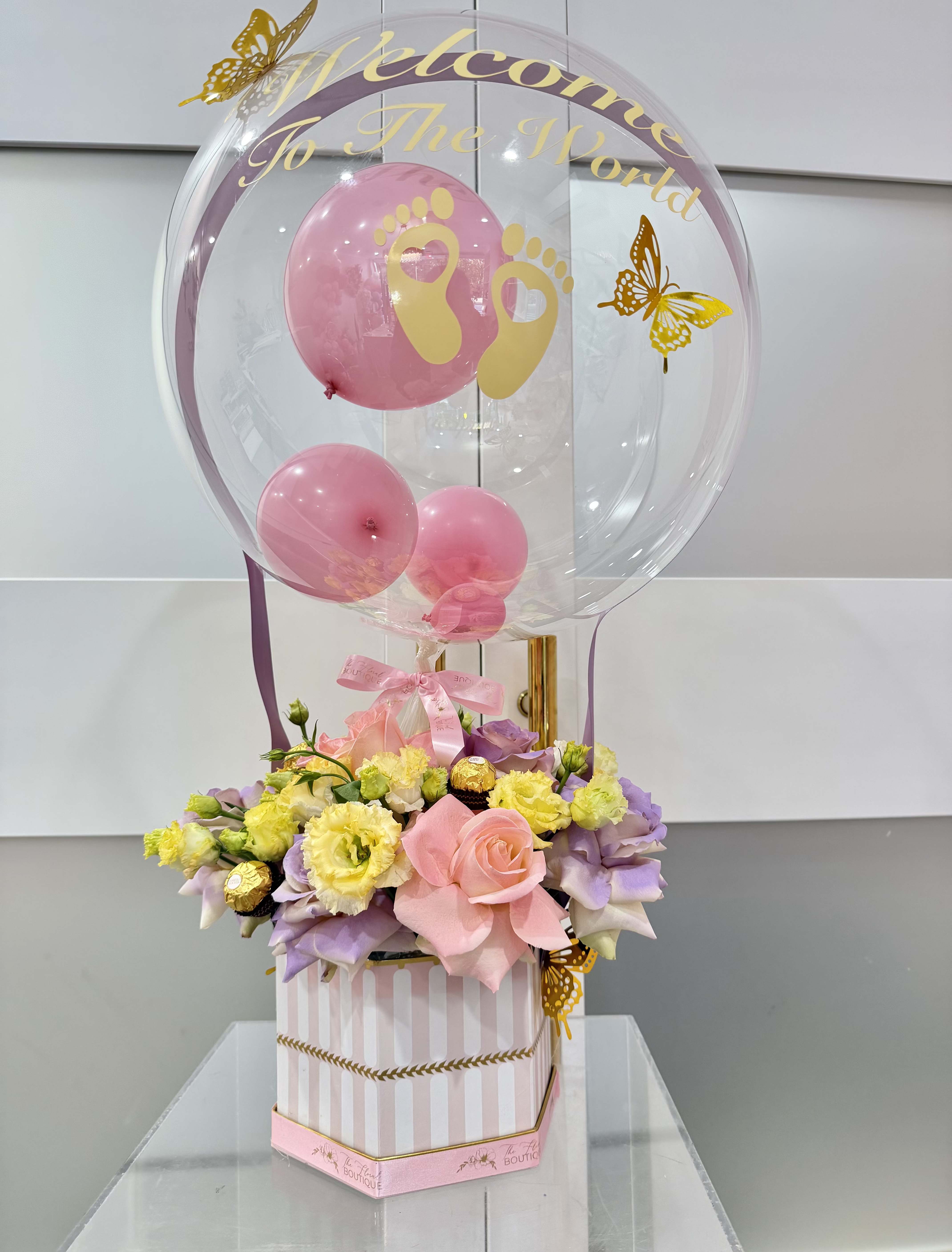  Welcome to the World Balloon Bloom Box  - Celebrate the arrival of a precious little one with our Welcome to the World Balloon Bloom Box! This charming arrangement features a large personalized bobo balloon adorned with delicate golden butterflies. Beneath the balloon, a lush mix of soft pink roses, pastel lavender blooms, it is beautifully arranged in a signature box. Touches of Ferrero Rocher chocolates add a hint of indulgence, making it the perfect gift set to welcome a newborn baby with love, sweetness, and style.   Perfect for: Baby showers • Newborn celebrations • Hospital or home welcomes  *** Let us know in the notes section if it's for a boy or girl.  ** Colors can be customized upon request.