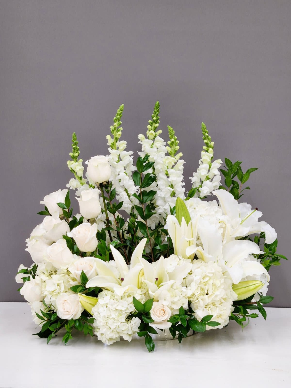 Peaceful Soul - Peaceful Soul is a serene cremation arrangement crafted with pure white snapdragons, roses, and lilies, accented by delicate spray roses and lush greenery. This elegant tribute honors a life well-lived with timeless blooms that convey grace, purity, and eternal peace. Perfect for expressing your deepest condolences during a difficult time.  Approximate Size:  Standard: 12" by 12"  Deluxe: 14" by 14"  Premium as Pictured: 18' by 22"