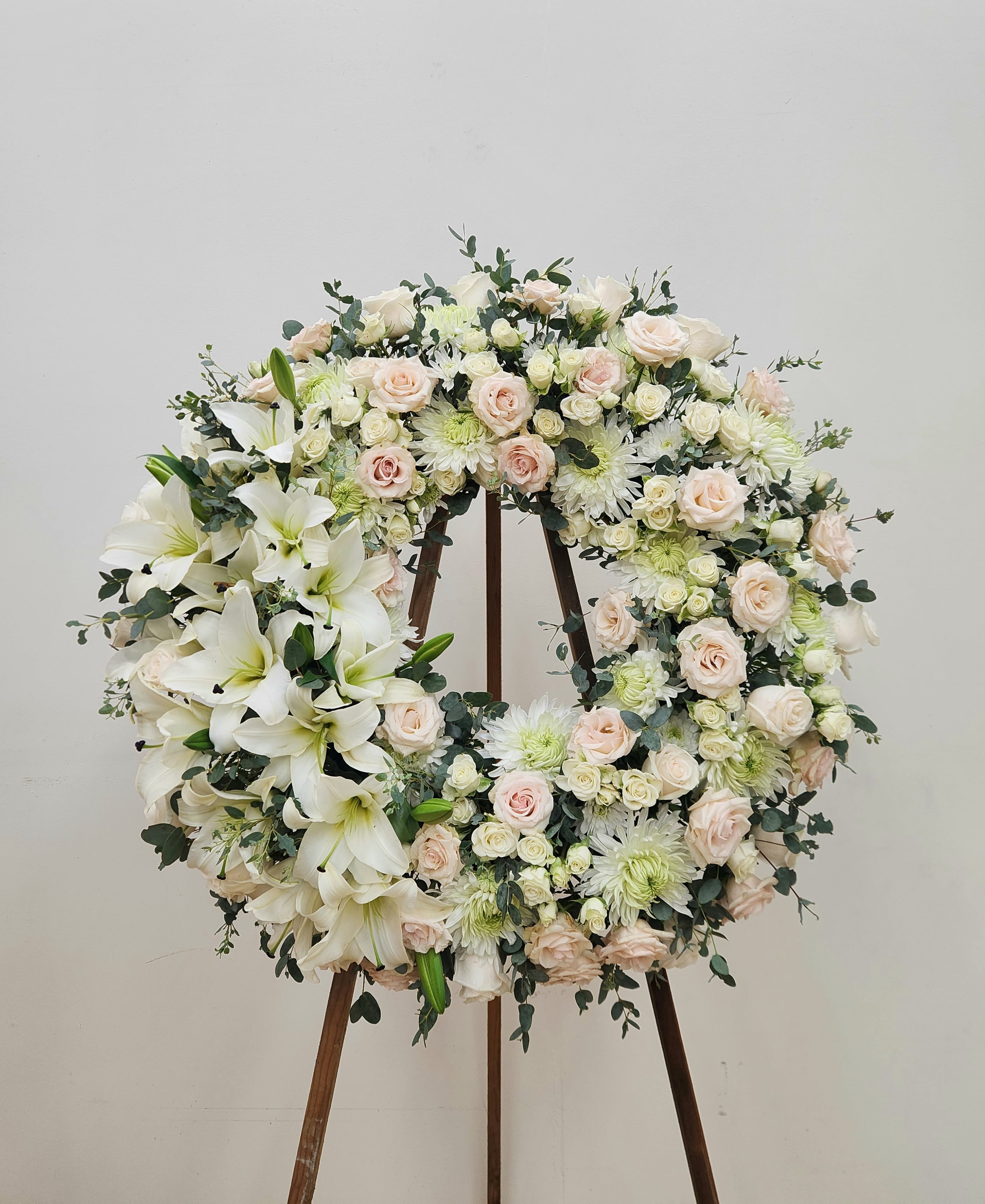 Meaningful Life Wreath - Honor a cherished memory with this elegant memorial wreath. Pristine white roses, mums, and lilies form the foundation, beautifully accented with soft pink spray roses and sage green baby eucalyptus that cascade around the entire piece. The lush, organic arrangement creates a refined yet naturally wild aesthetic—a sophisticated tribute that speaks to a life well-lived. Perfect for services, celebrations of life, or heartfelt remembrance.
