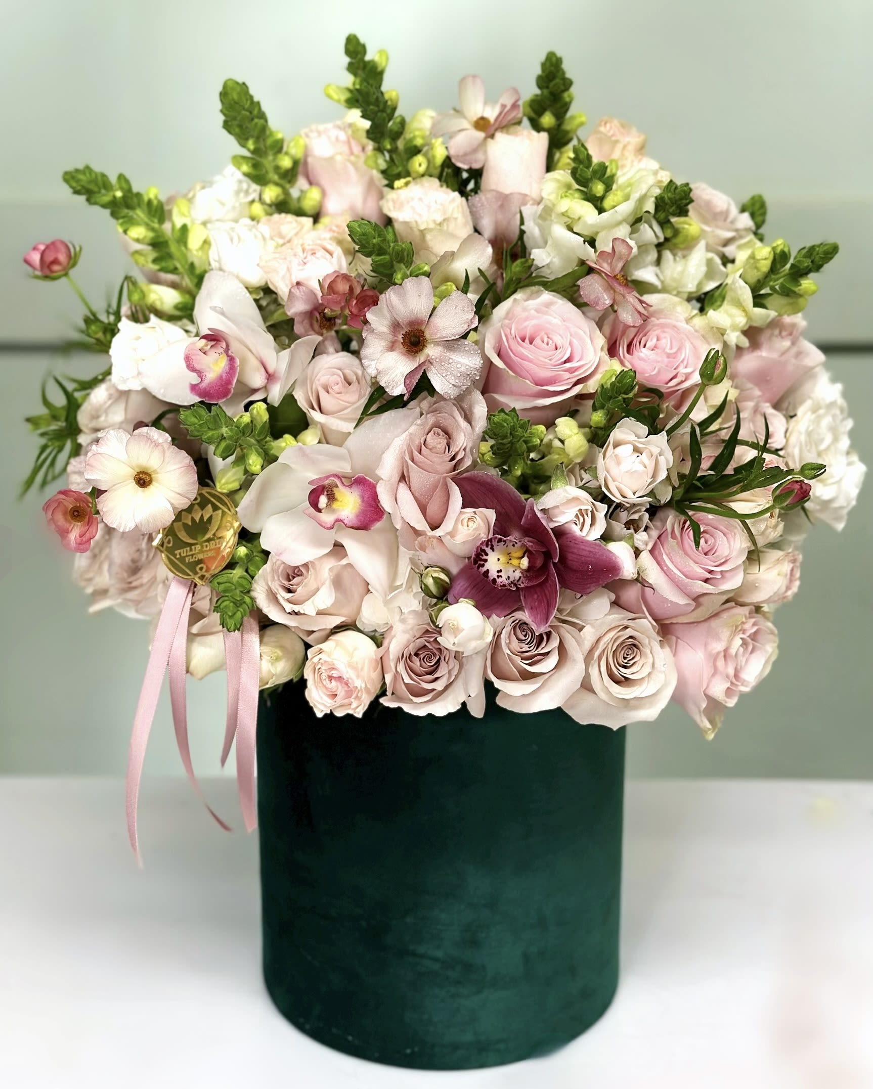 Blushing Elegance Box  - A romantic blend of pale pink roses, orchids, lisianthus, and ranunculus, this lush design in a velvet green box is made to melt hearts. Perfect for a graceful and elegant Valentine’s Day surprise. Please note: The image shown represents our Premium Size arrangement.