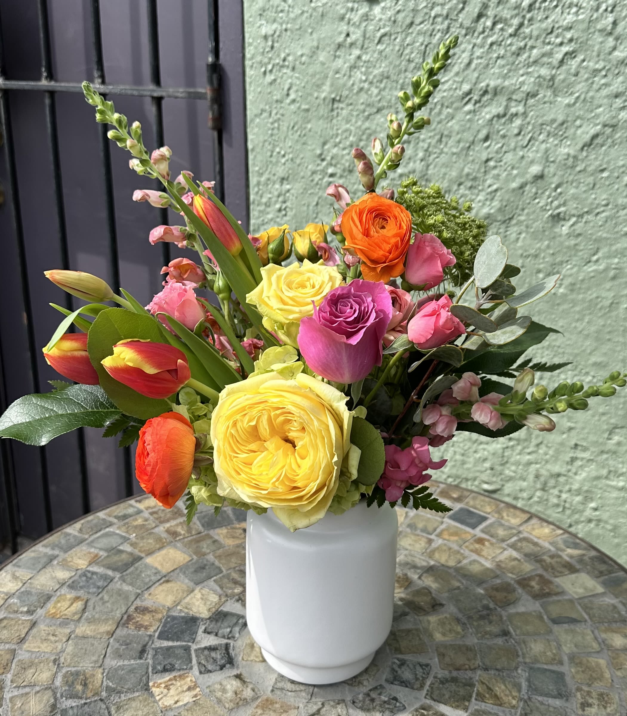 Color Pop - A vibrant and joyful burst of color designed to uplift any space, this arrangement is a celebration of nature's most radiant summer blooms. Featuring a lush mix of bright pink garden roses, sunny yellow dahlias, and towering orange snapdragons, it brings the warmth and energy of a golden afternoon indoors. Handcrafted with a variety of textures and hues, this arrangement offers a little piece of sunshine, perfect for making any occasion special or simply brightening someone's day.