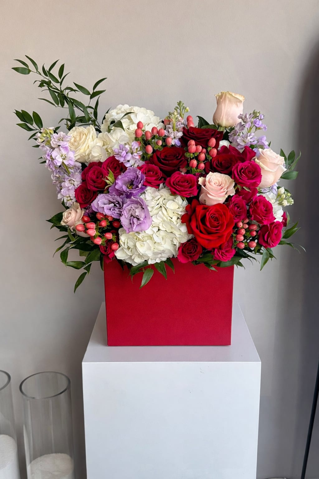 Lush arrangement of red and pink roses with white hydrangeas and purple blooms in a red square box