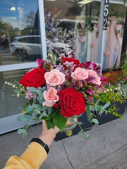 Small vase arrangement of red and pink roses with accent flowers, held in front of a shop window