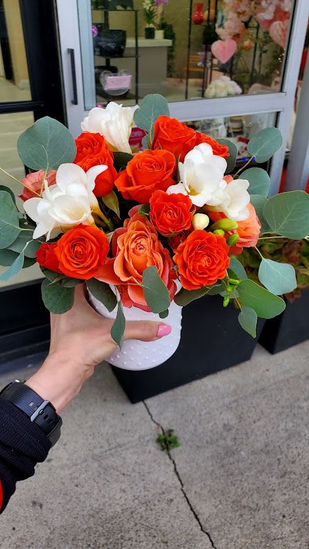 Compact orange and white rose arrangement in a white pot held outside a flower shop