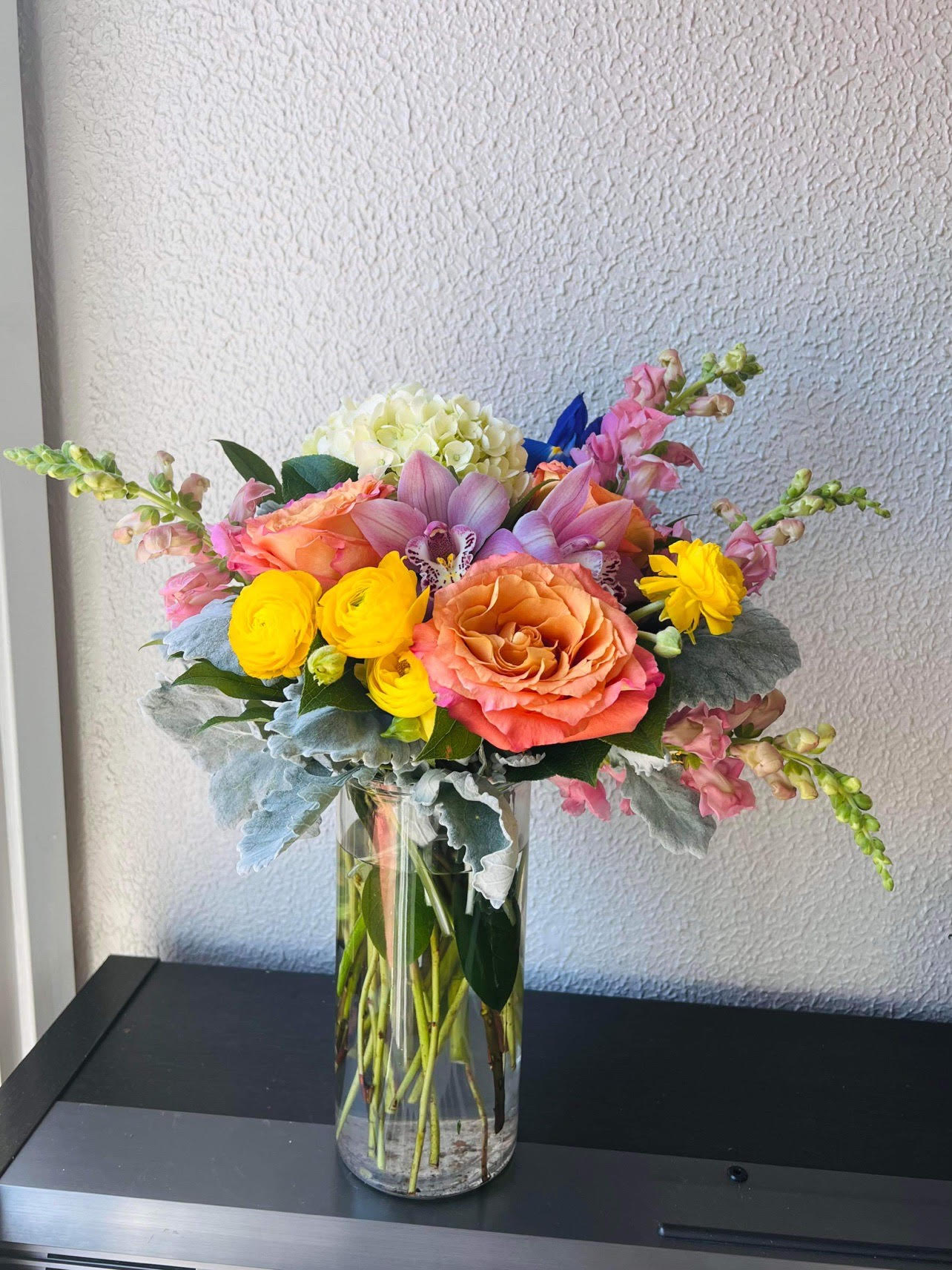 House of Blossoms - - 4 Pink Snapdragons - 3 Orange Roses - 4 Yellow Ranunculus - 2 Pink Cymbidium Orchids - 1 White Hydrangea - Cylinder Glass Vase