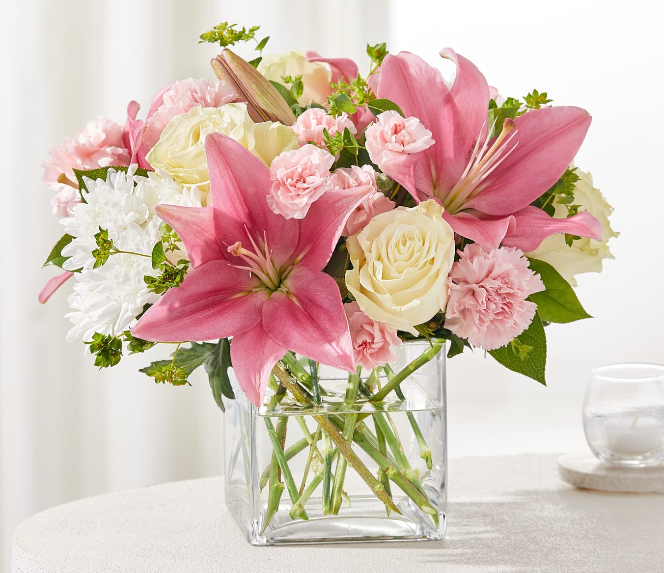 By Your Side Bouquet - Show your love and support with this soothing blend of blooms. White roses and soft pink lilies are paired with white mums, carnations, and lush greenery, creating a gentle expression of care and comfort.  Designed in a modern cube vase, this bouquet is a beautiful way to remind someone they’re never alone.  Approximately 12" H x 14" W