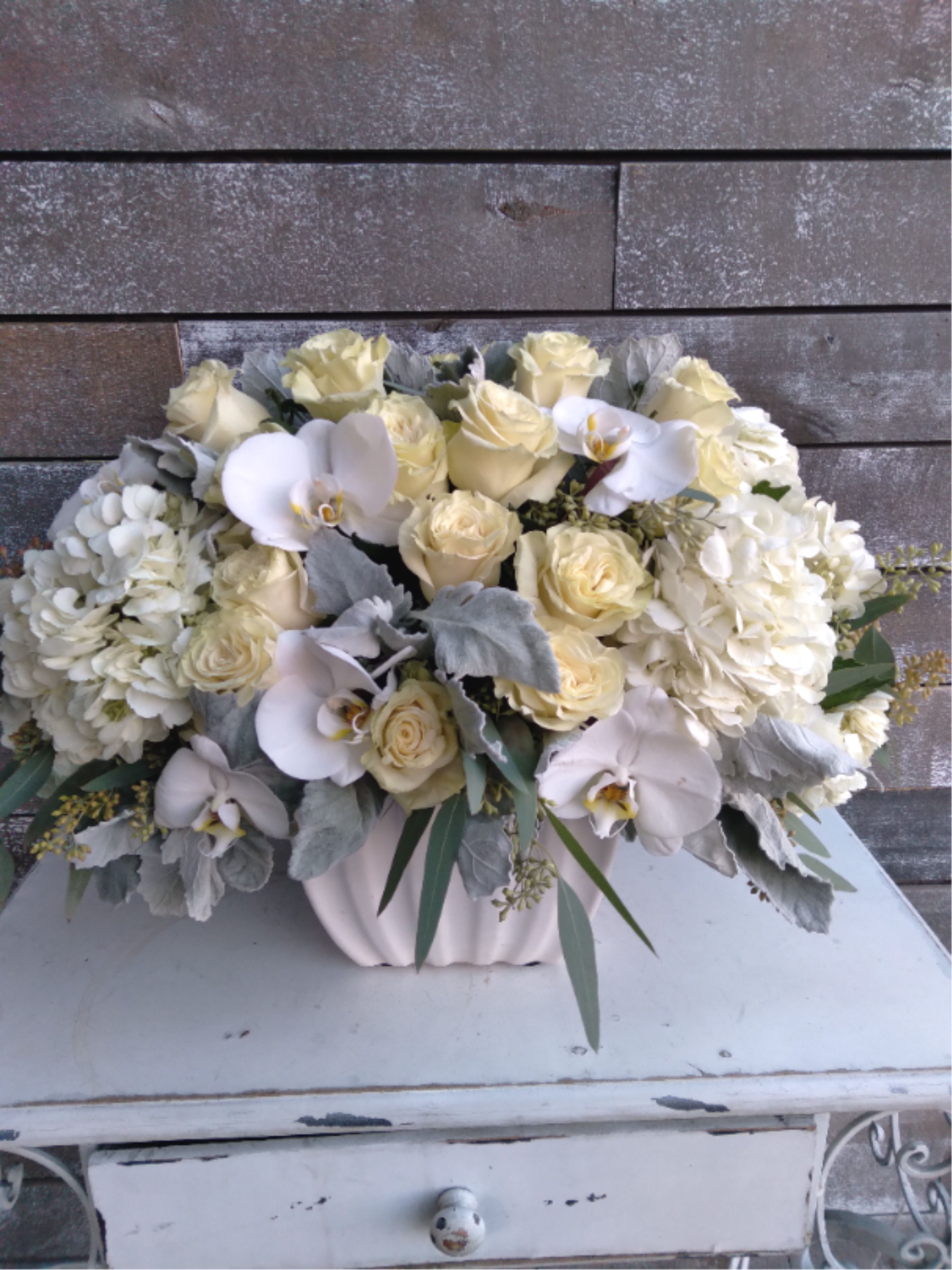 Ivory Elegance - Ivory Elegance is a luxurious composition featuring creamy ivory roses, lush white hydrangeas, and graceful white Phalaenopsis orchids. Accented with soft silver foliage and delicate greenery, this arrangement creates a harmonious palette of warmth, texture, and sophistication.  Designed in a sculptural ivory vessel, the composition balances fullness with softness, offering a romantic yet structured presence. The blend of premium blooms and muted tones evokes understated luxury and classic elegance — never overpowering, always refined.