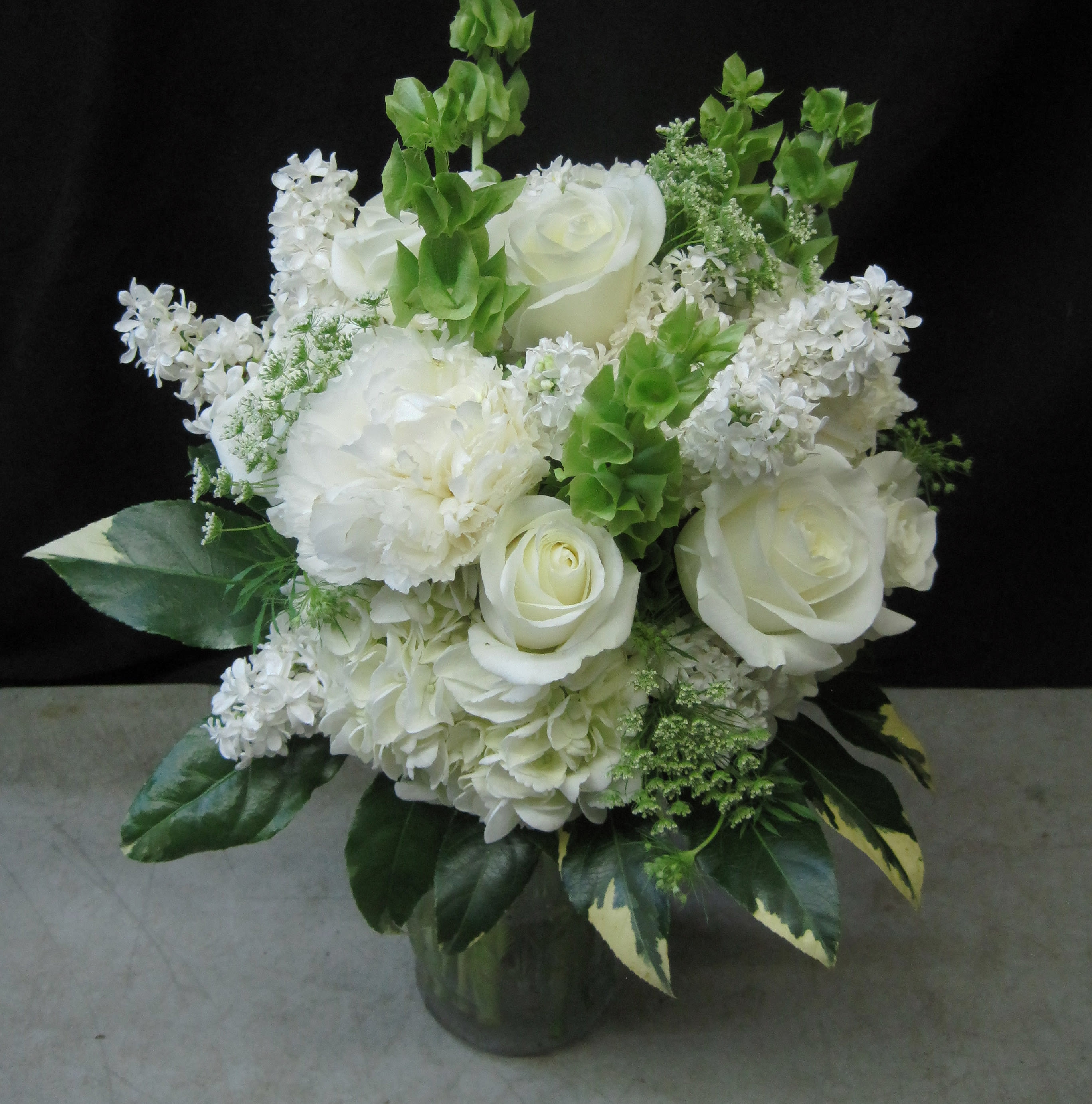 Simply  White - Clean and elegant in purest white, this timeless look suits so many occasions and  fits into any decor. It is featured here with Roses, Carnations, Hydrangea, Queen Ann's Lace, Bells of Ireland and variegated green and white foliage. 
