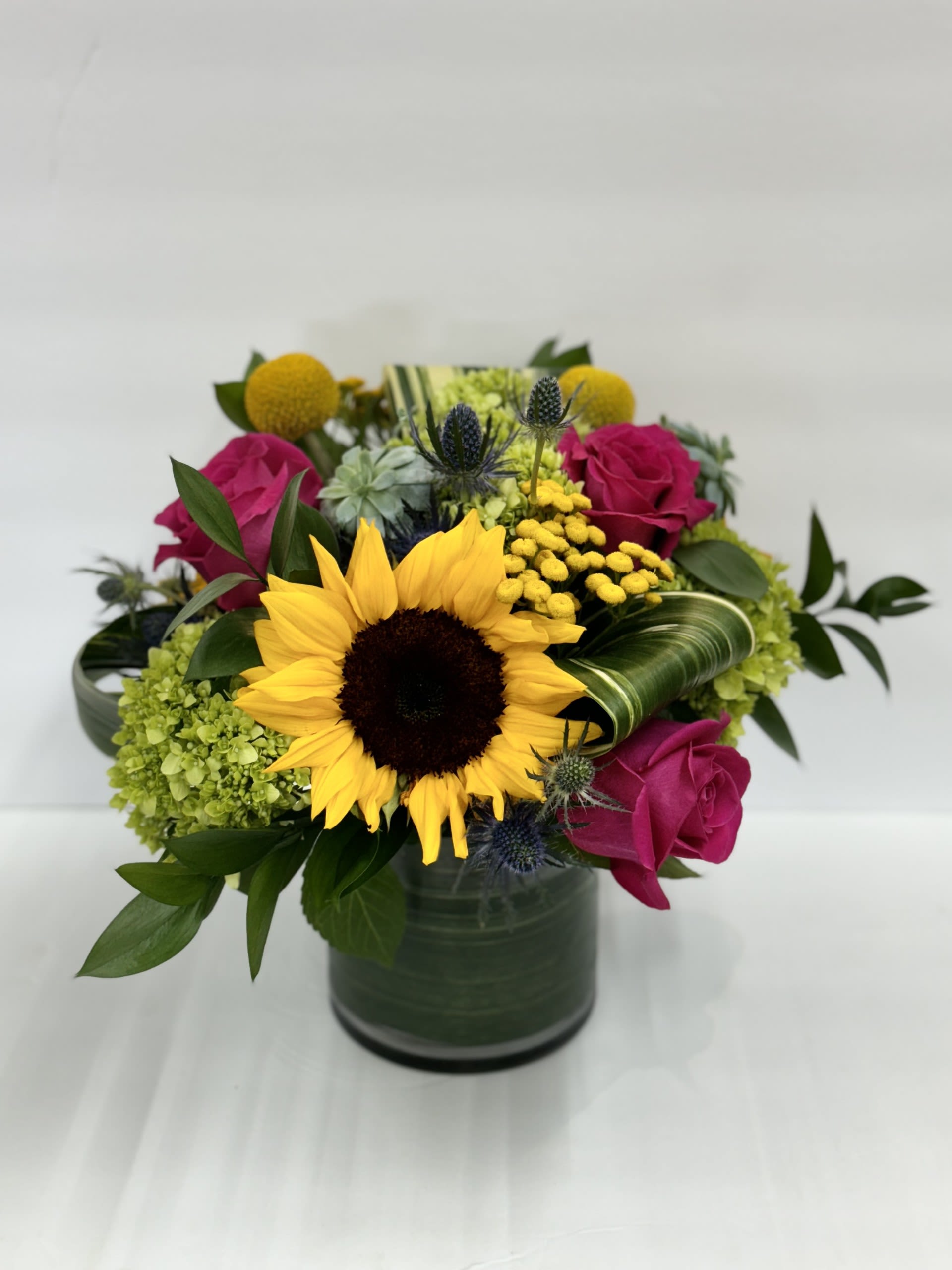 Timeless Sunset Bouquet - Celebrate your special someone's with the "Timeless Sunset Bouquet." Whether it's a mile stone birthday or congratulating your loved one, this vibrant arrangement will be the perfect gift to put a smile on their face and brighten up the space. Sunflowers, Hot Pink Roses, Green Hydrangeas, and Bill Bobs are the focal flowers nestled in greenery.  