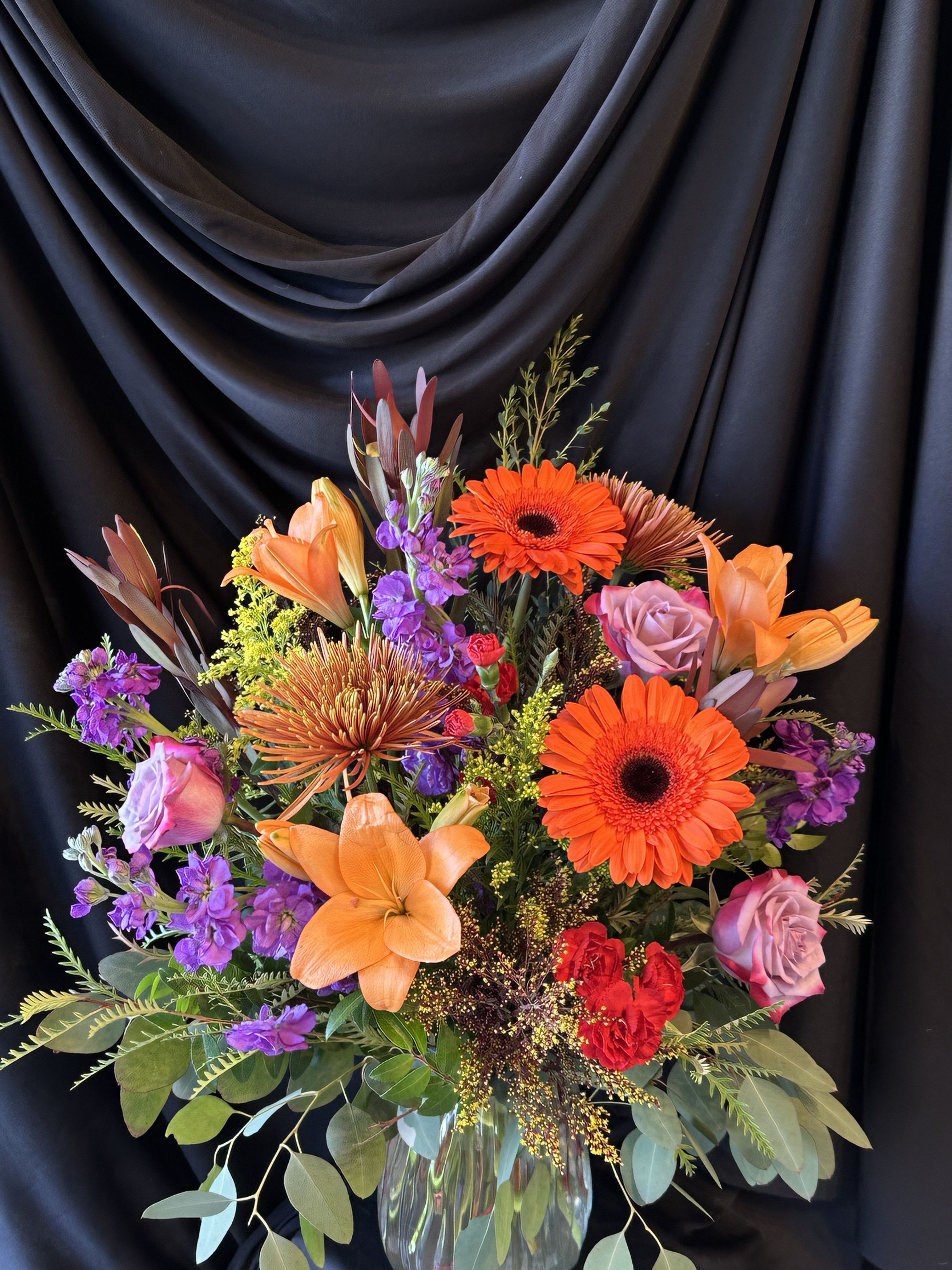 Bright mixed bouquet with orange lilies, gerbera daisies, and purple roses in a glass vase