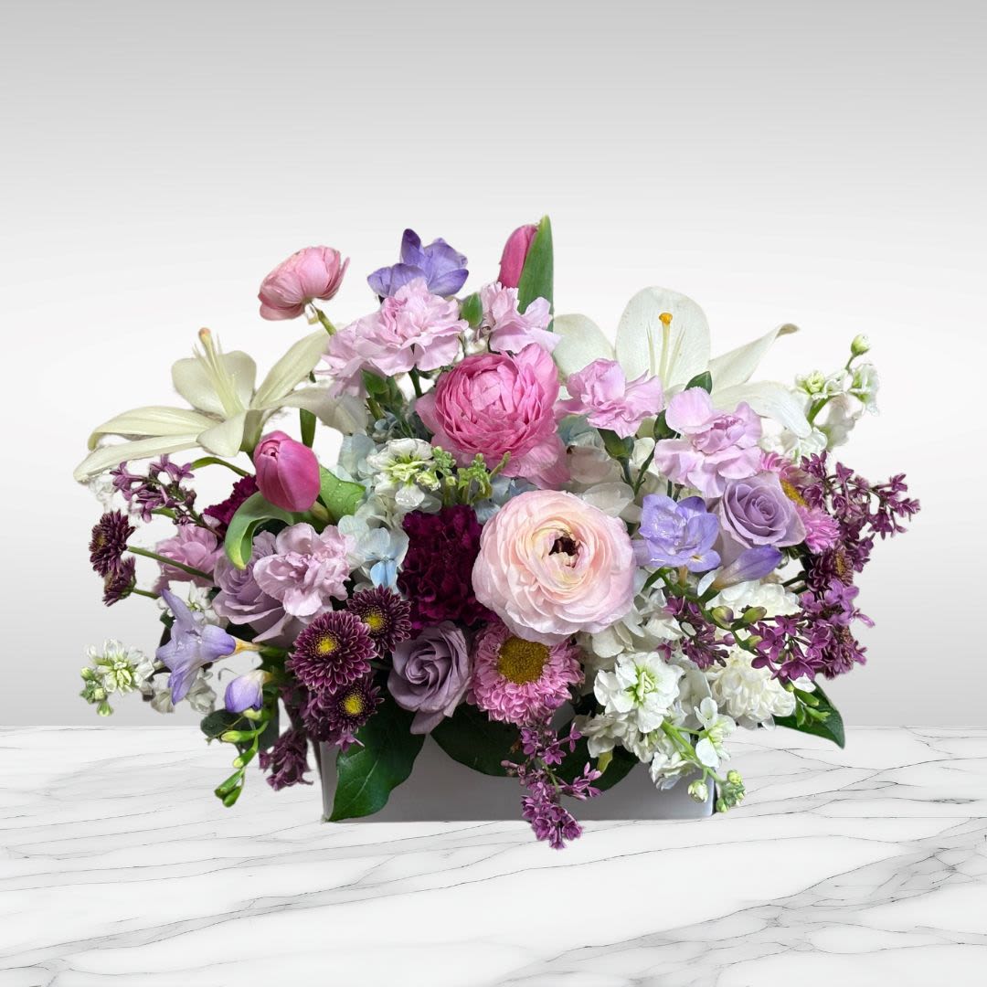 Bountiful Spring Blossoms - Recipe: White Lilies, White Stock, Seasonal Ranunculus, Seasonal Llilac, Freesia, Lavender Roses, Carnations, Matsumoto Aster, Pink Tulips, and more in a white rectangular, ceramic vase. Seasonal: Seasonal Ranunculus, Lilac Orientation: Front Facing View Photo shown: Standard