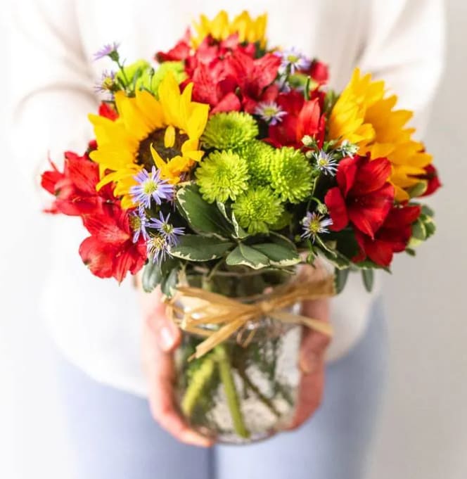 Fields of Flowers - It's the beauty of a field of flowers from the comfort of home. This bouquet of flowers has a chic farmhouse style while still staying modern. Bright sunflowers surrounded by pink alstroemeria and green poms are displayed in a mason jar wrapped in raffia. The essence of a field of flowers can be brought to anyone's home, and will charm anyone who sees it!